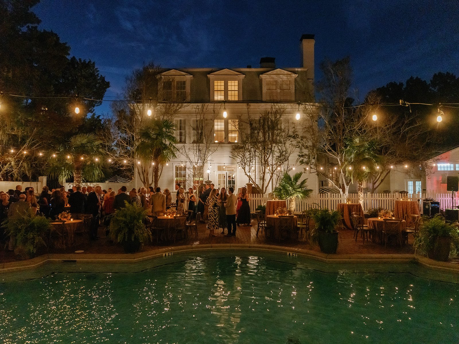 Evening poolside wedding welcome party with guests mingling beneath string lights in front of a historic white house.