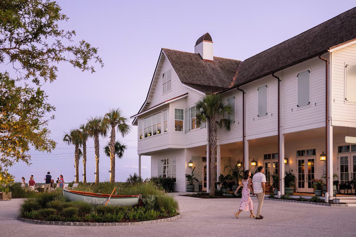 The Dunlin exterior at dusk with a white coastal building, palm trees, warm porch lights, and guests walking near the entrance and lawn.