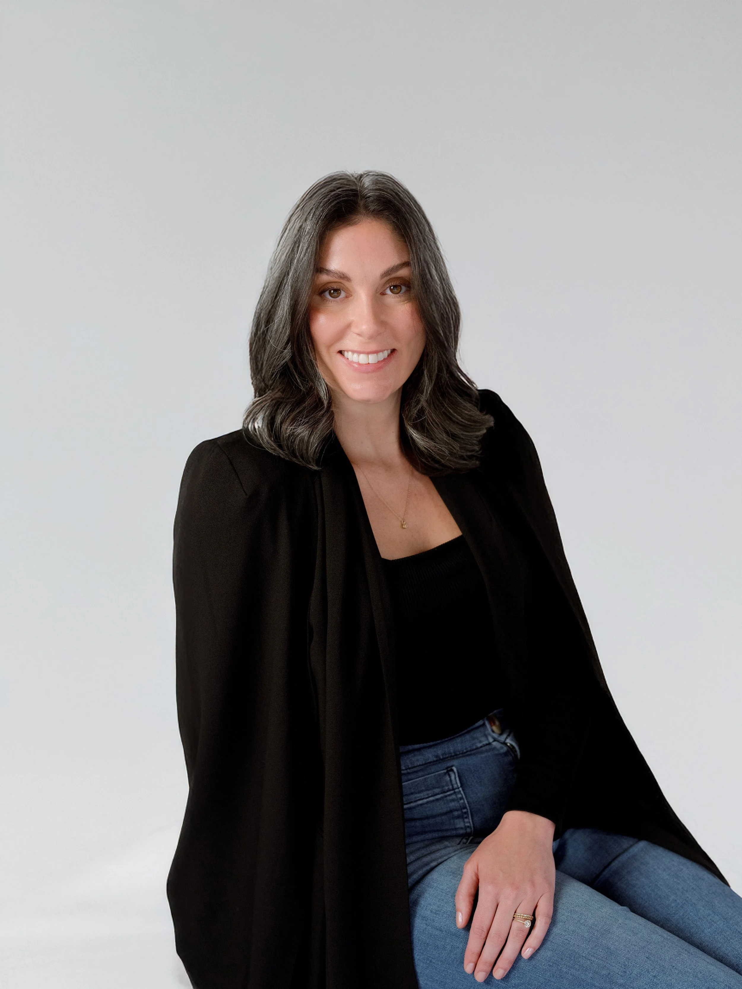 Portrait of an EventDox co-founder seated against a soft gray studio background, wearing a black blazer, black top, and blue jeans.