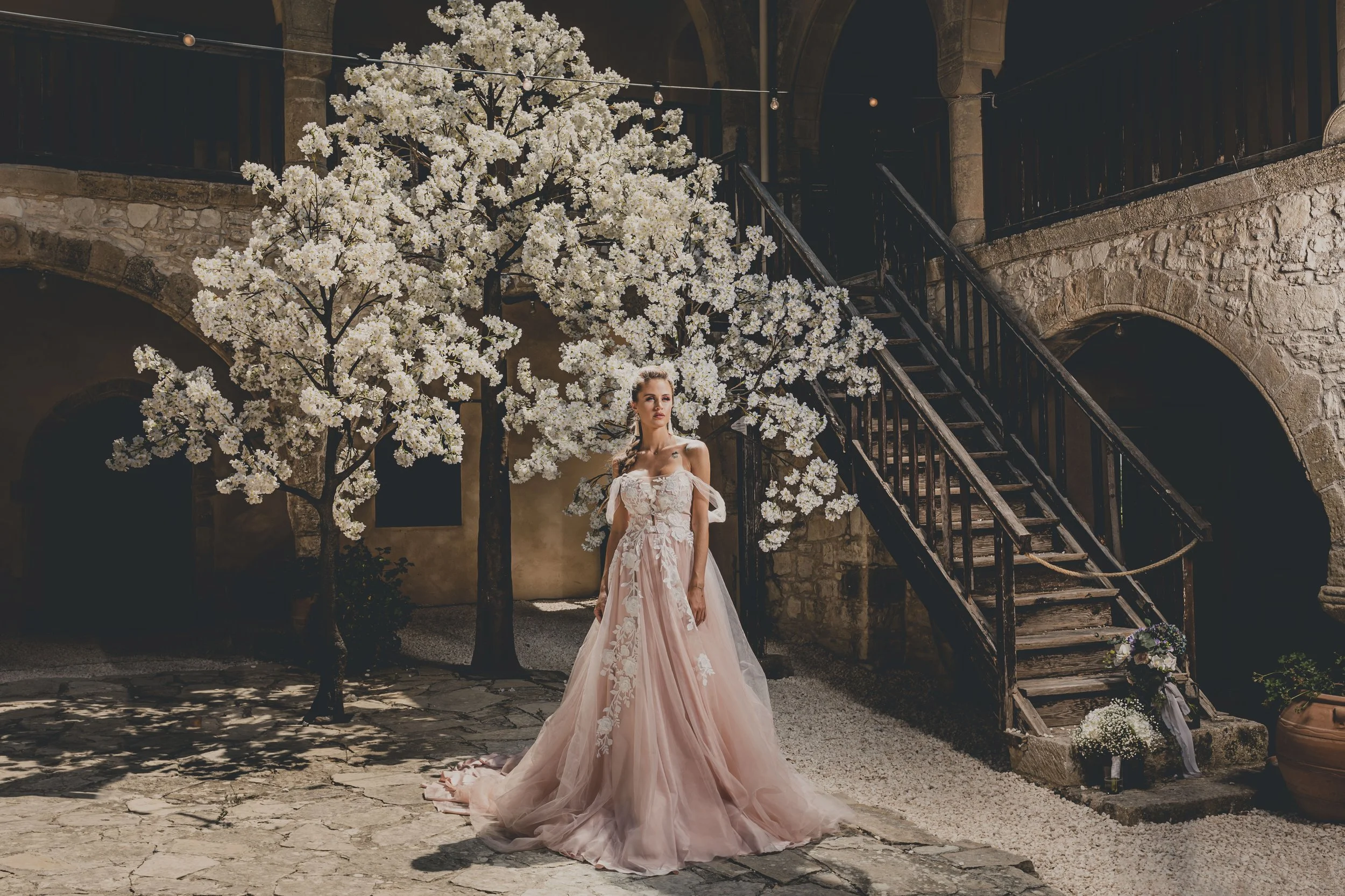 Bride in a blush gown standing in a stone courtyard beneath blooming white trees, framed by rustic architecture and a wooden staircase.