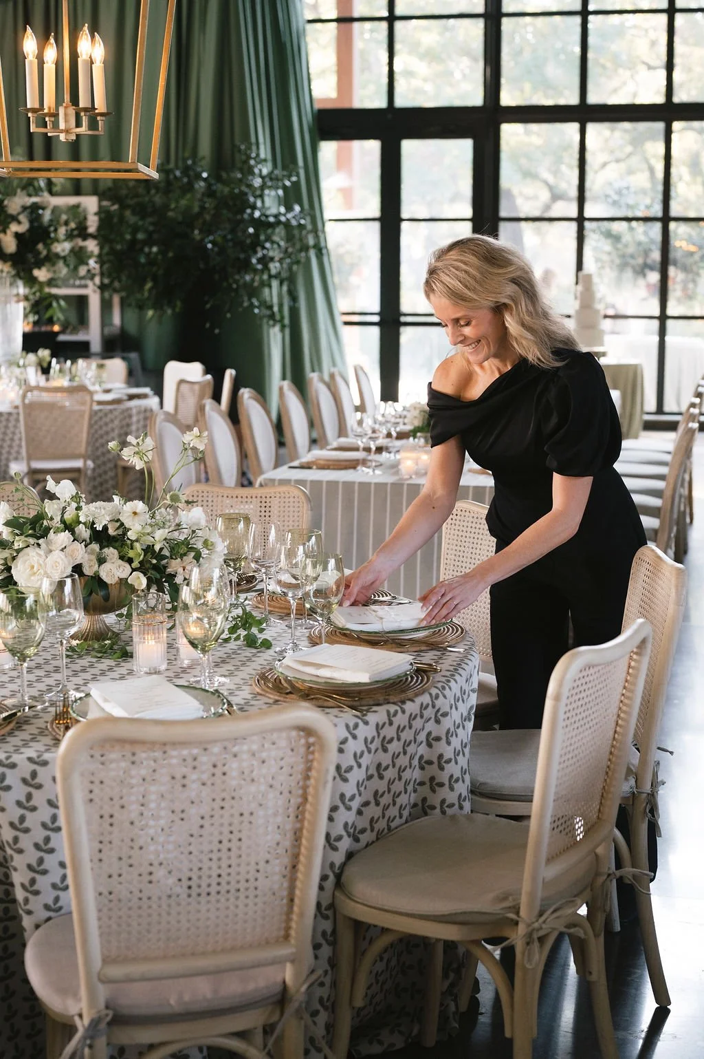 Event planner arranging a wedding reception place setting beside patterned linens, white florals, glassware, and woven dining chairs.