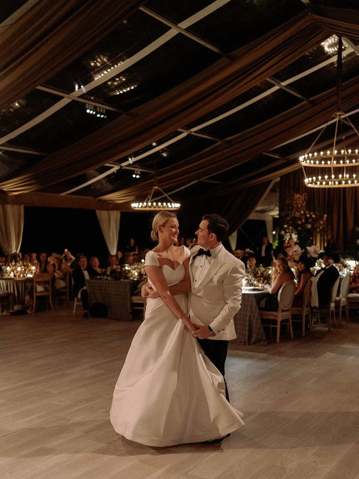 A bride and groom share their first dance beneath a softly lit tented reception, surrounded by seated guests and warm candlelight.