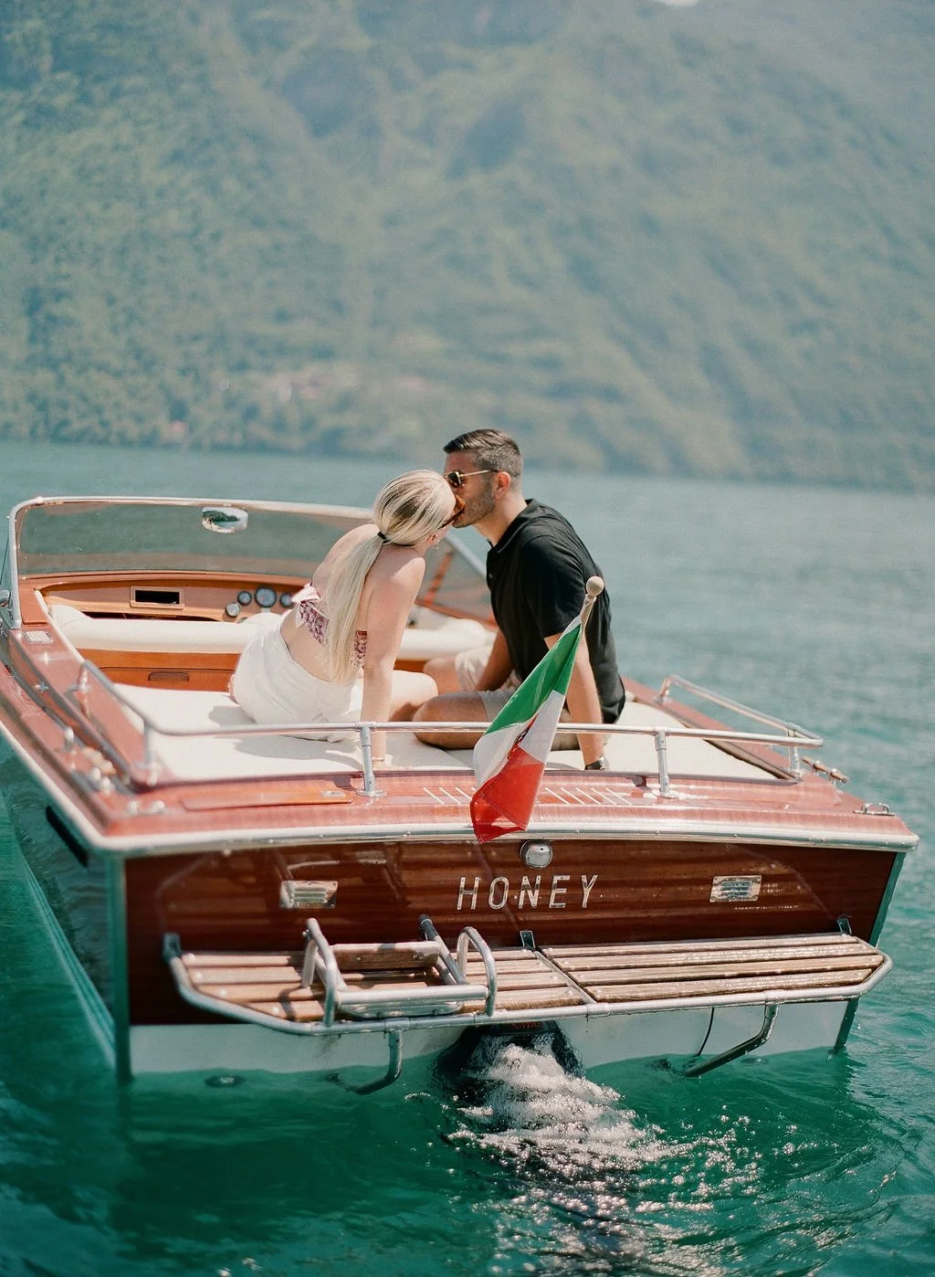 Couple kissing on a classic wooden boat on Lake Como with an Italian flag and mountain views in the background.