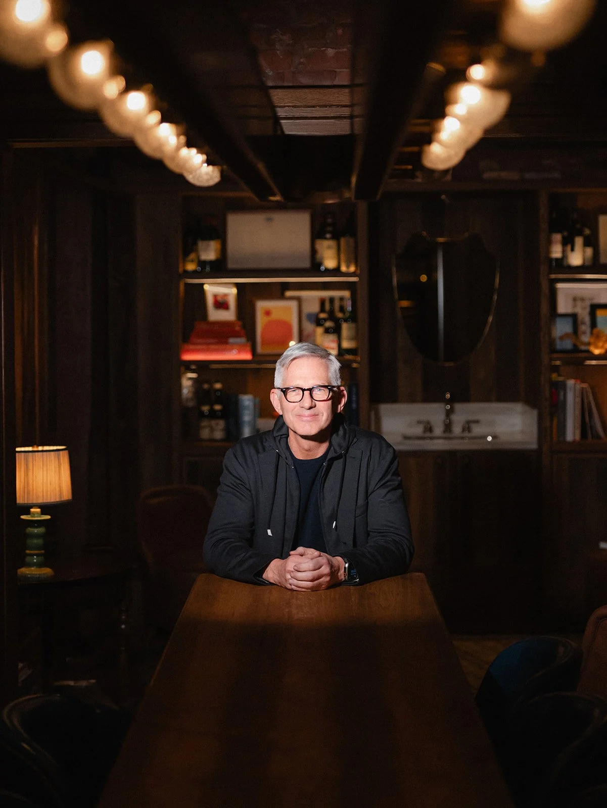 Bryan Rafanelli seated at a long wooden table in a dark, warmly lit interior with shelves, lamps, and wine bottles in the background.