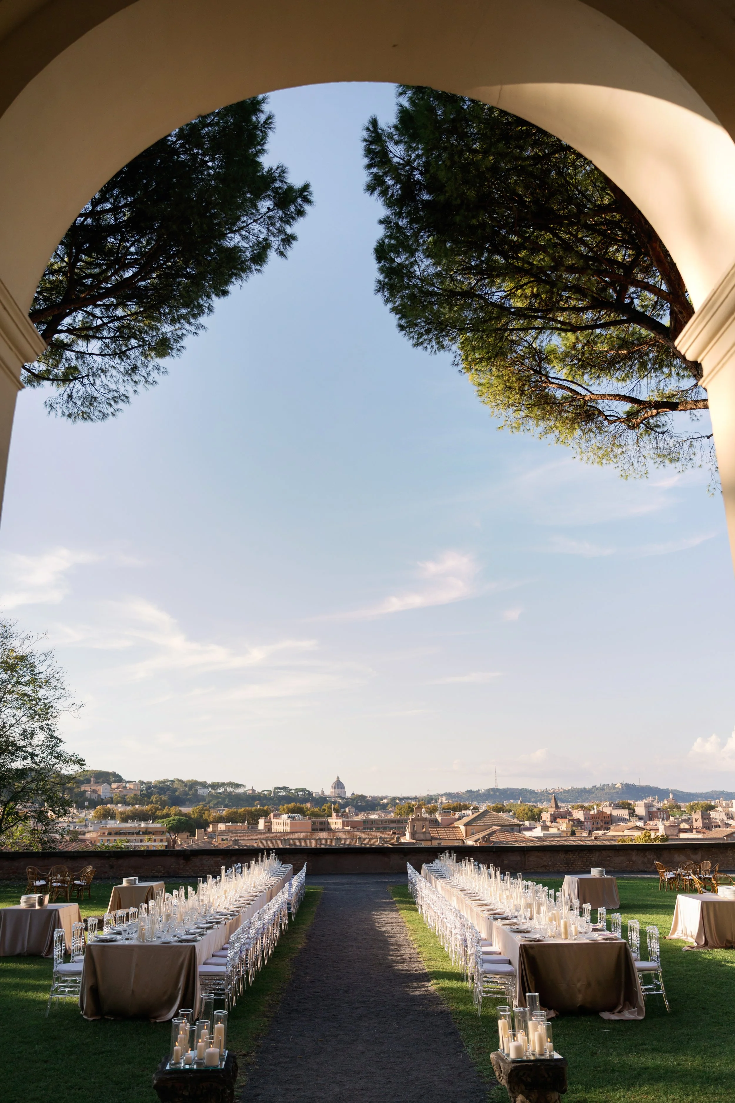 Outdoor banquet tables set beneath an archway with a sweeping view toward the Rome skyline and soft evening light.