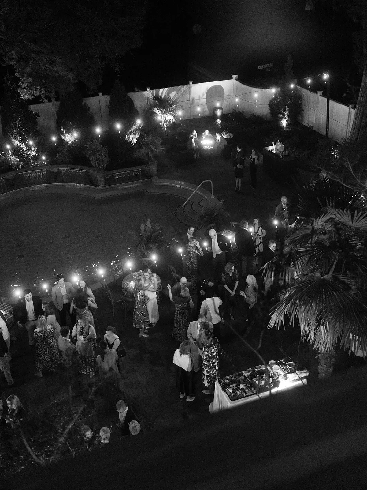 Evening wedding weekend gathering around a pool with string lights, cocktail tables, lush landscaping, and guests mingling outdoors.