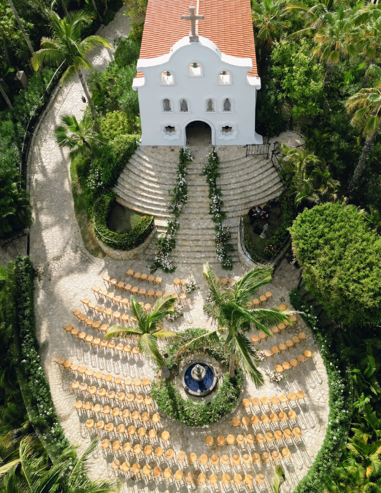 Aerial view of a white chapel with a terracotta roof surrounded by palms, with ceremony seating arranged in rows and a fountain in the center.