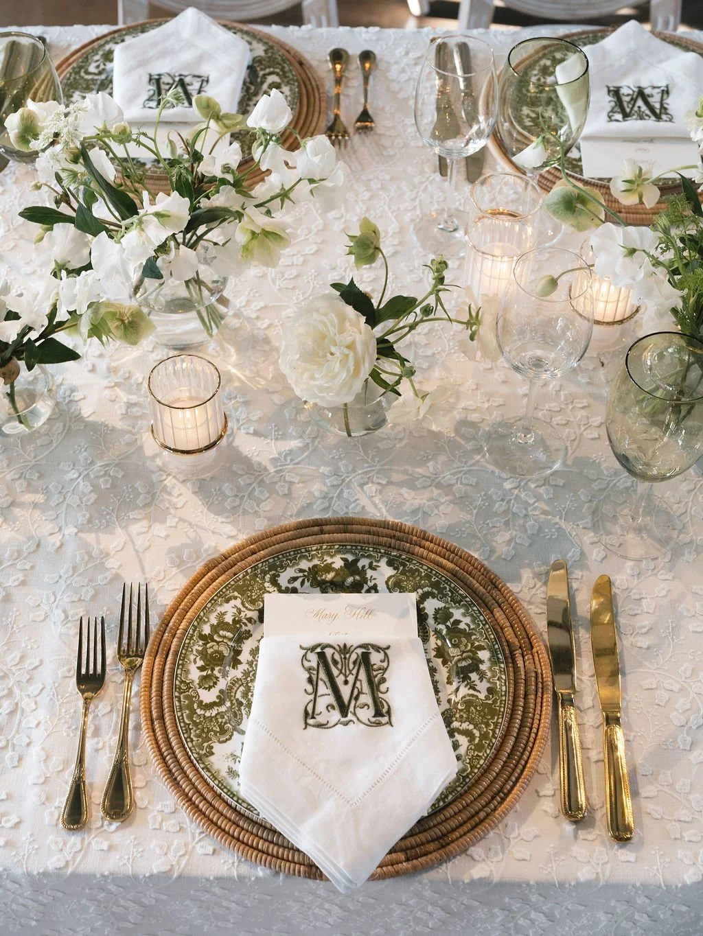 Detailed reception place setting featuring green patterned china, woven charger, gold flatware, and embroidered monogram napkin surrounded by soft white florals and candlelight, styled by Reagan Events.