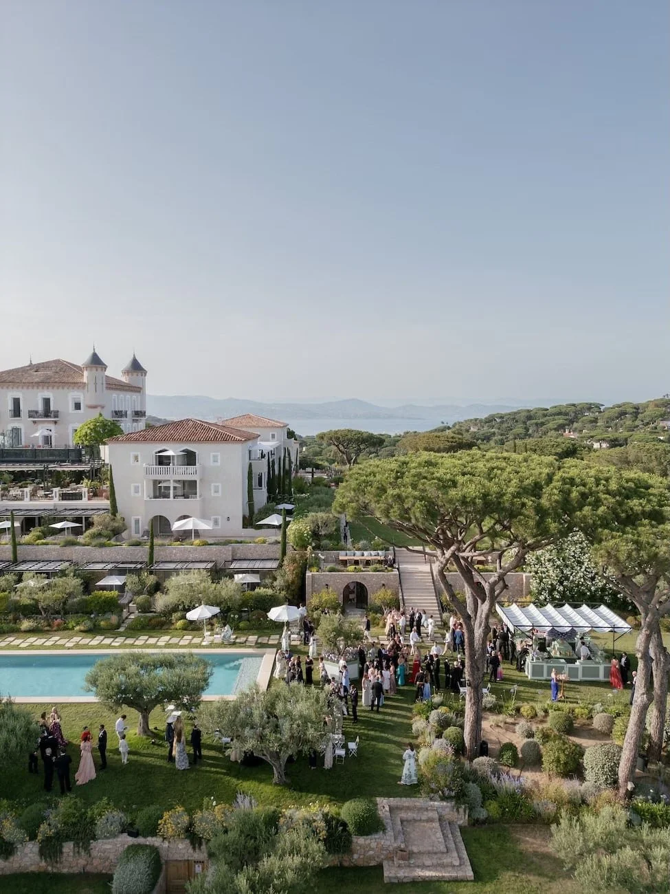 Aerial view of a coastal estate with terracotta roofs, a pool, and guests mingling on a garden lawn set for cocktail hour, with the sea in the distance.