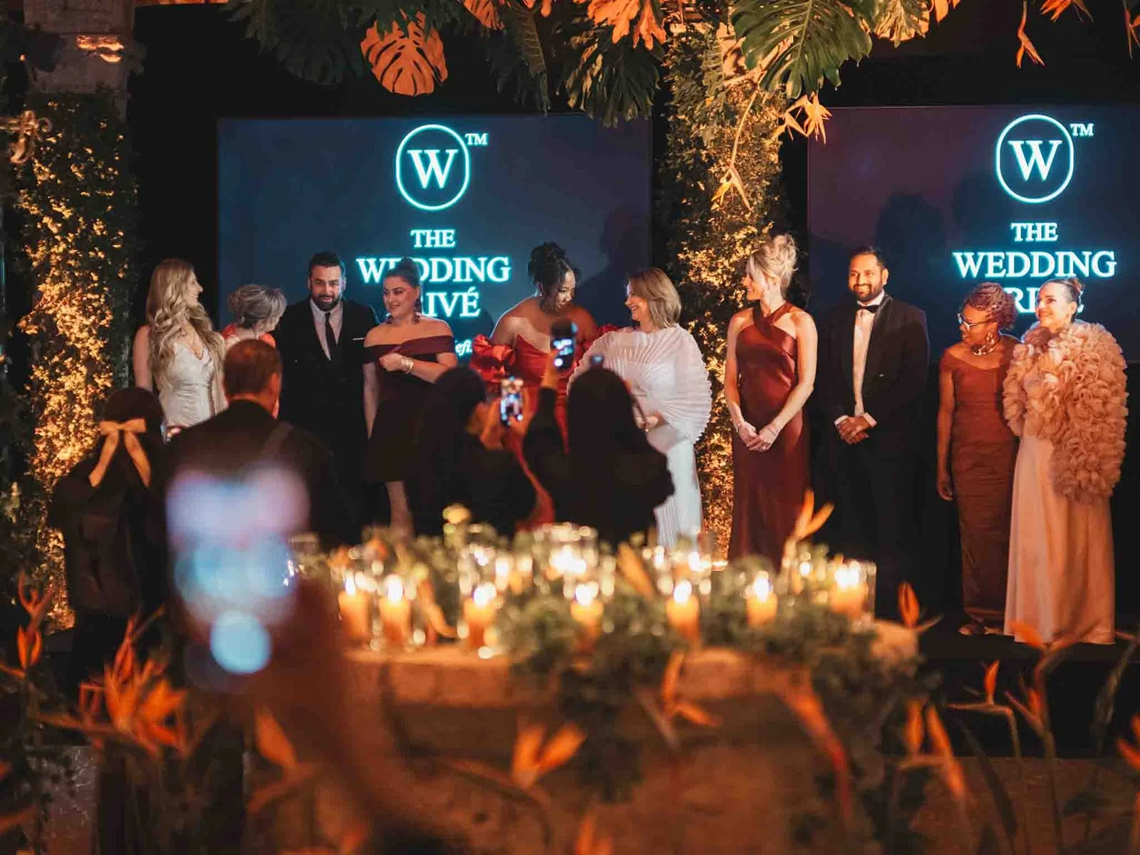 Members of The Wedding Privé gathered on stage during the Rome Première as guests watch from a candlelit reception setting.