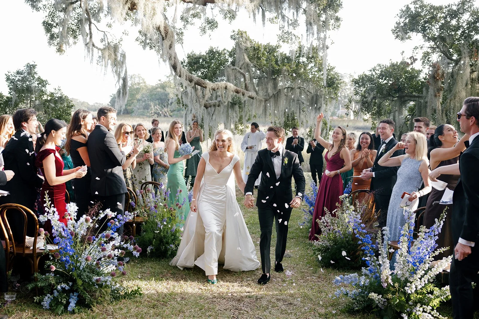 Couple stands with their officiant on a grassy oceanfront lawn as guests sit in two rows facing the ceremony, with dunes and the ocean behind them.