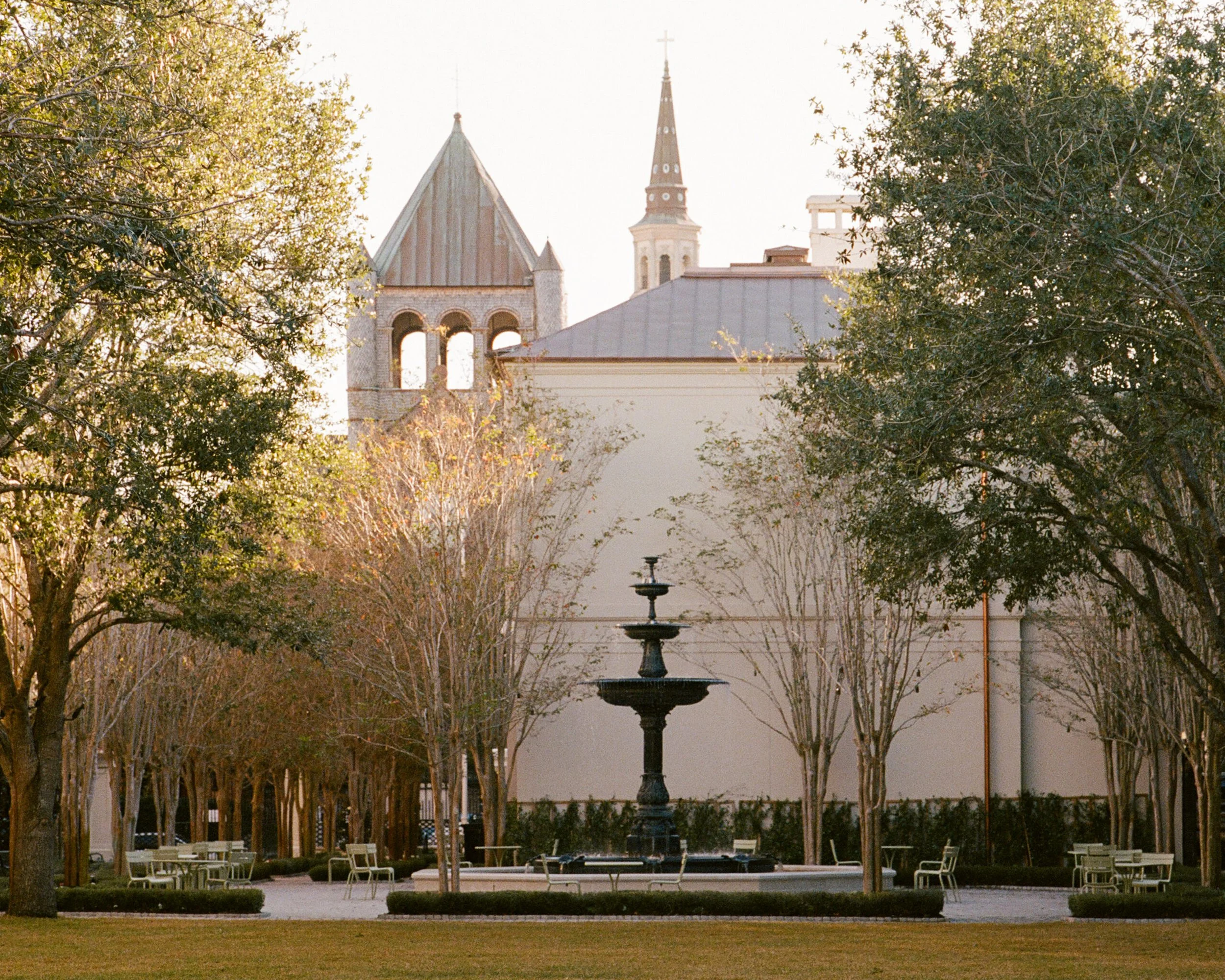 American Gardens in downtown Charleston with a tiered fountain, tree-lined lawn, garden seating, and historic church architecture in the background.