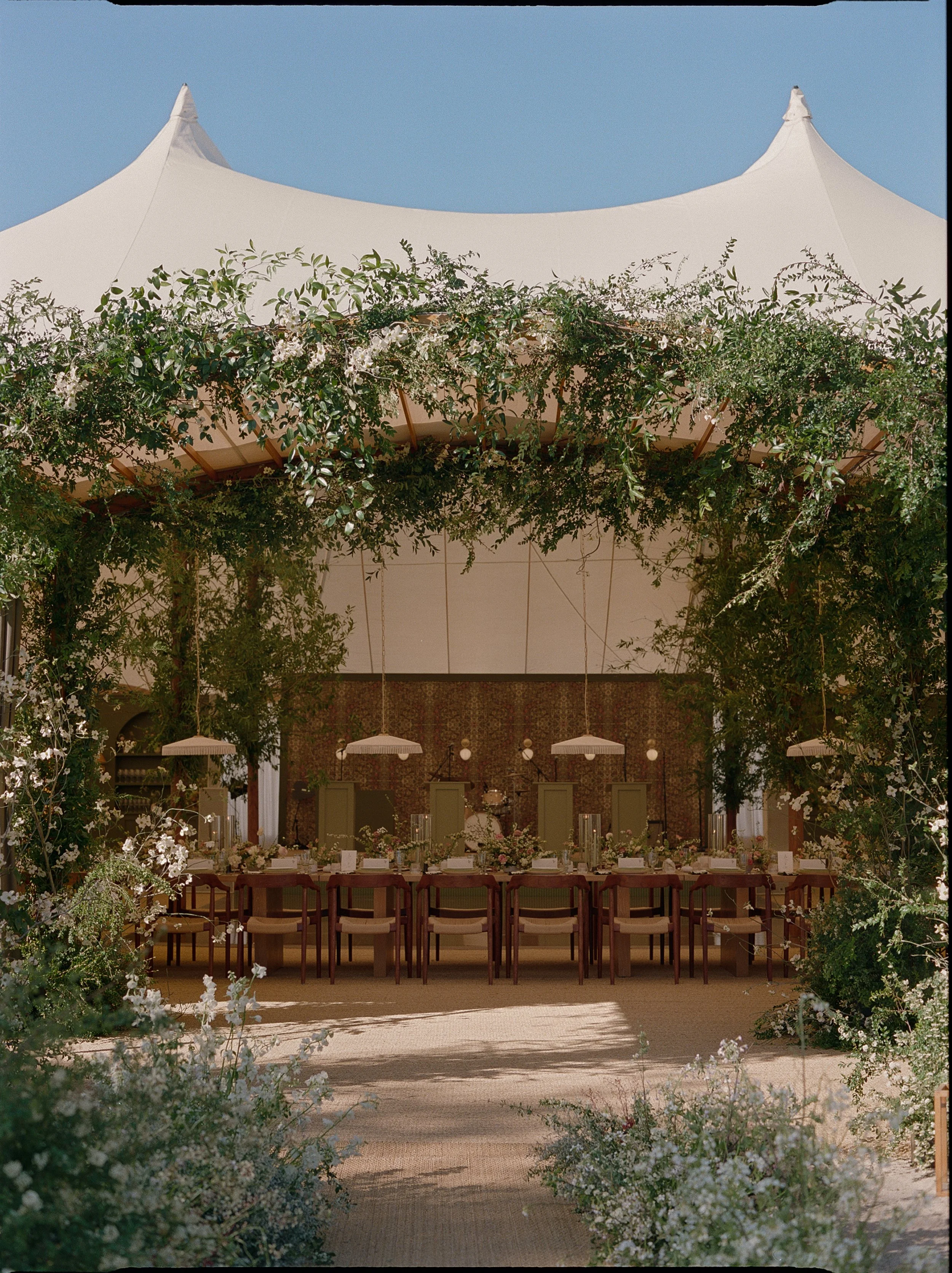 Open-air tented wedding reception with a long dining table framed by abundant greenery, white flowers, and suspended lighting.