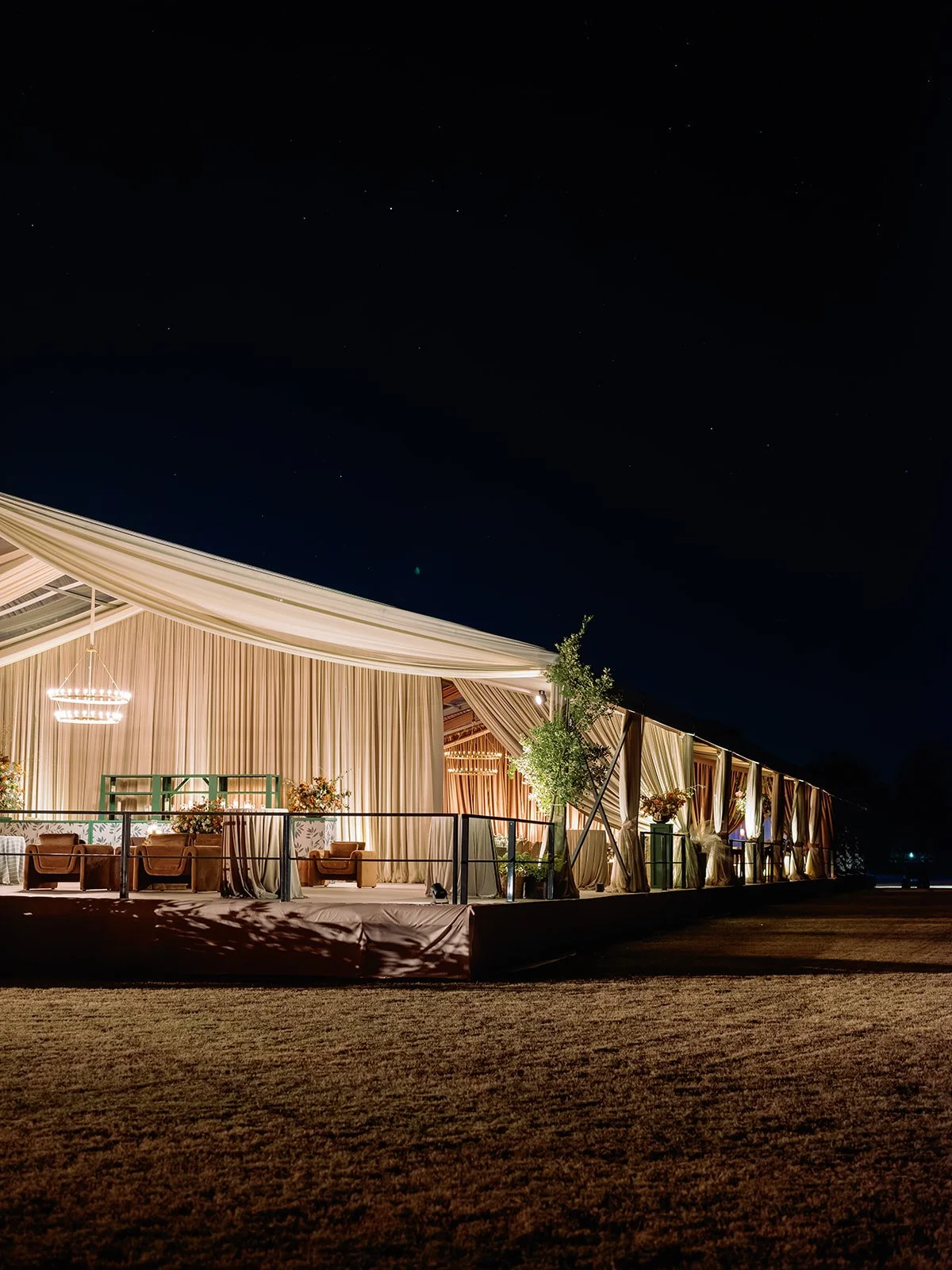 Exterior view of a tented wedding reception at night with draped entrances, chandeliers, and lounge seating glowing under the evening sky.