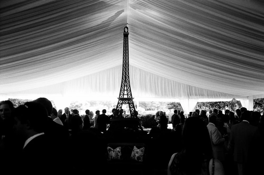 Black-and-white view of wedding guests beneath a fully draped reception tent with an Eiffel Tower installation as the focal point.