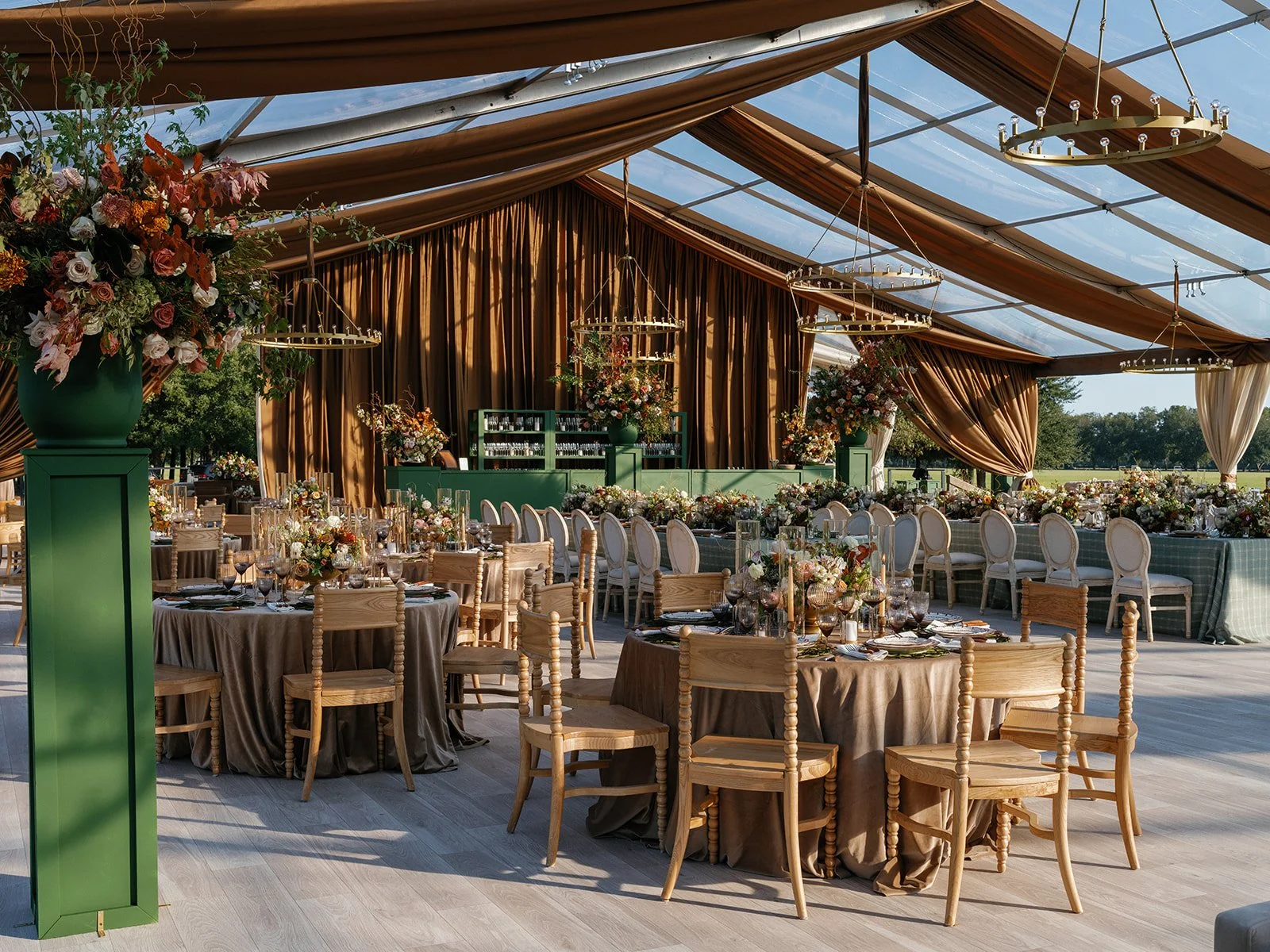 Tented wedding reception with brown ceiling draping, green bars, abundant floral arrangements, round dinner tables, and open views to the surrounding landscape.