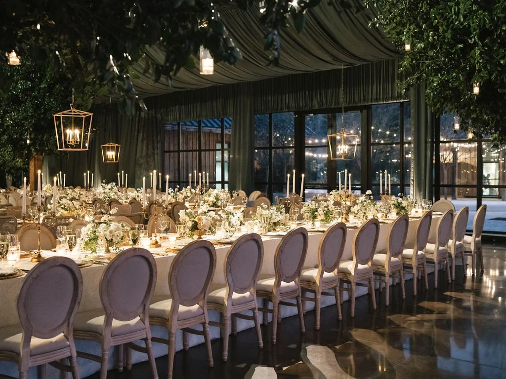 Candlelit wedding reception with long tables, white florals, and a draped ceiling inside a glass-walled event space at The Dunlin.