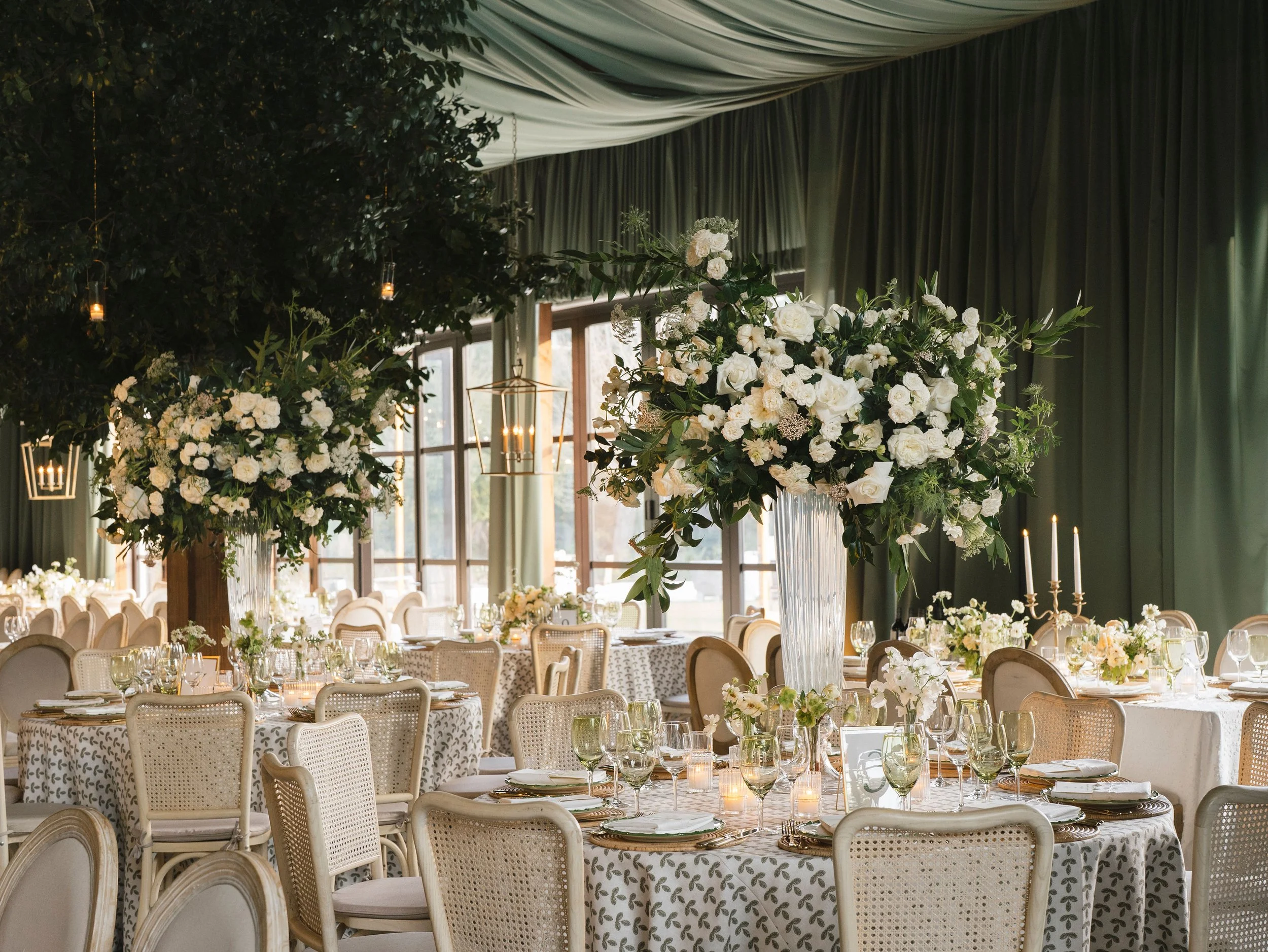 Reagan Events wedding reception with oversized white floral arrangements, green draping, candlelit tables, and cane-back dining chairs.