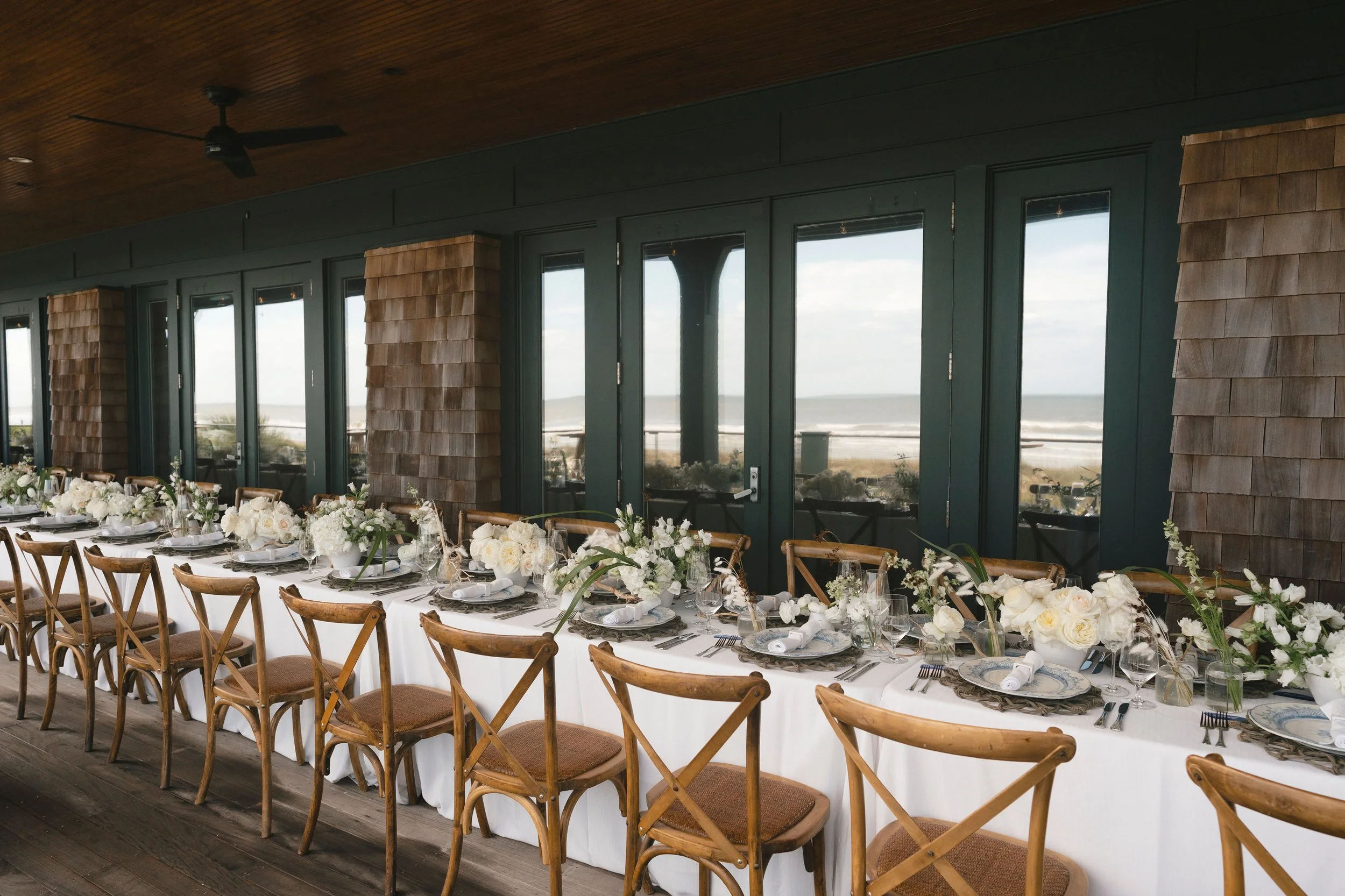 Long dinner table set with white linens, natural wood chairs, and low white floral arrangements on a covered porch with ocean views on Kiawah Island.