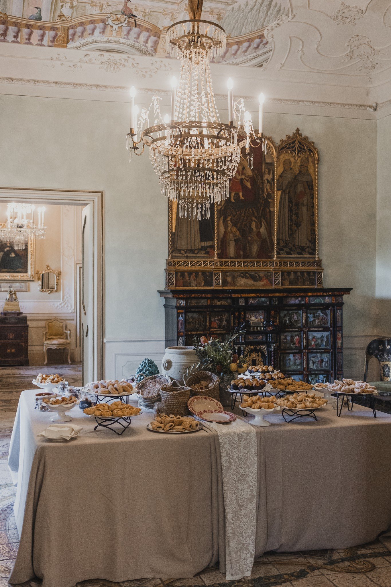 An ornate Italian villa salon with crystal chandeliers and historic artwork, featuring an abundant table of pastries and desserts for a wedding gathering.