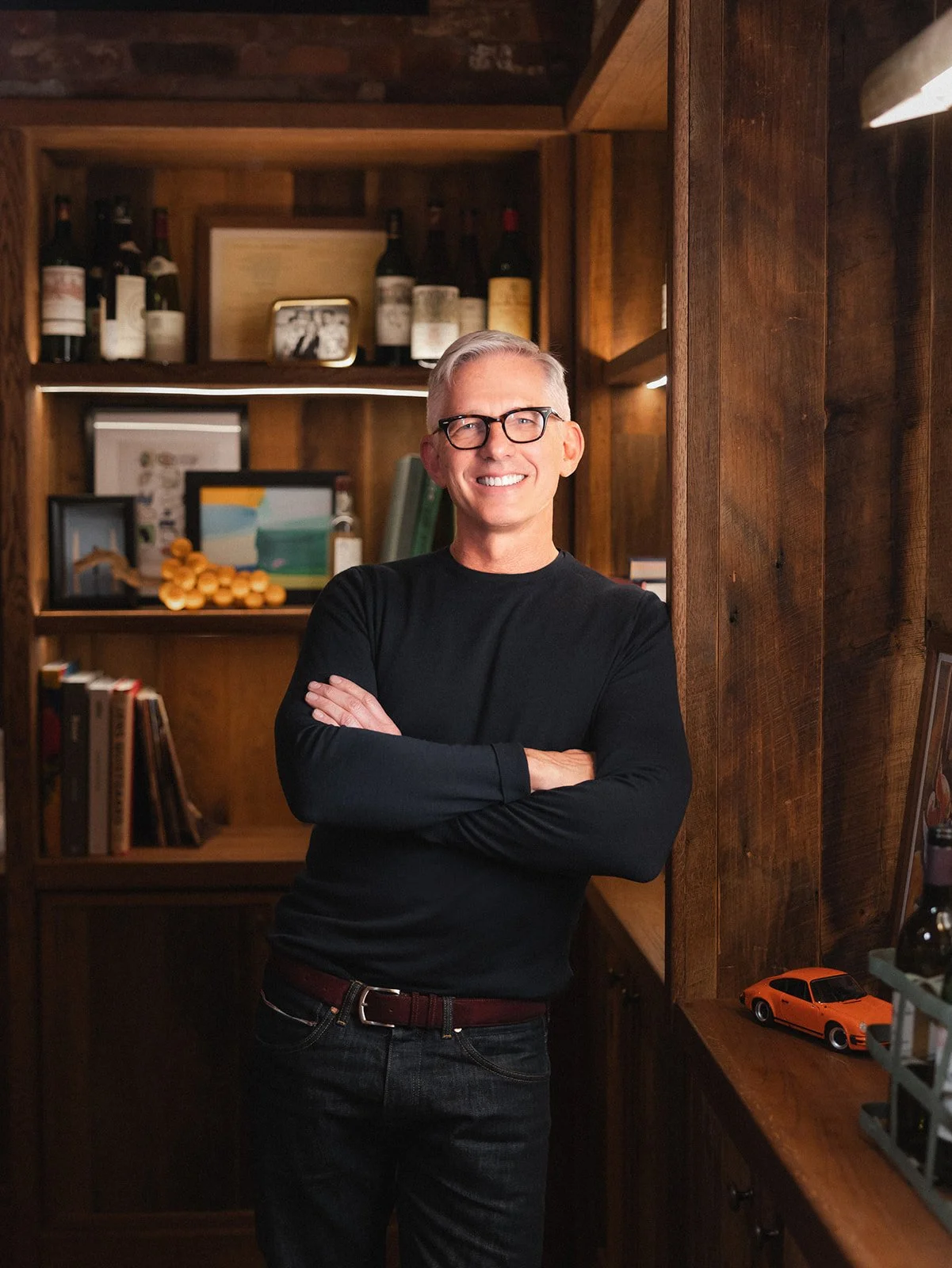 Bryan Rafanelli standing with arms crossed in a wood-paneled office interior with books, framed artwork, and warm shelving behind him.