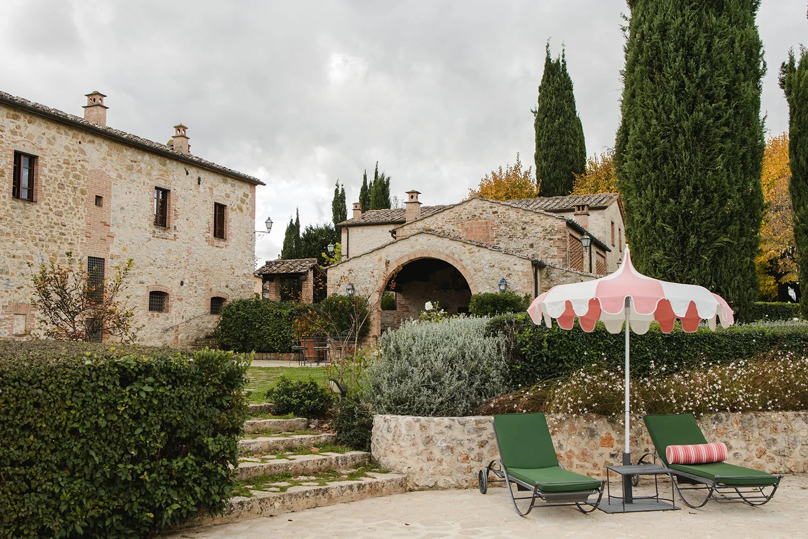 Stone borgo-style villa in Tuscany with cypress trees, garden paths, and green lounge chairs beside a pool with a pink striped umbrella.