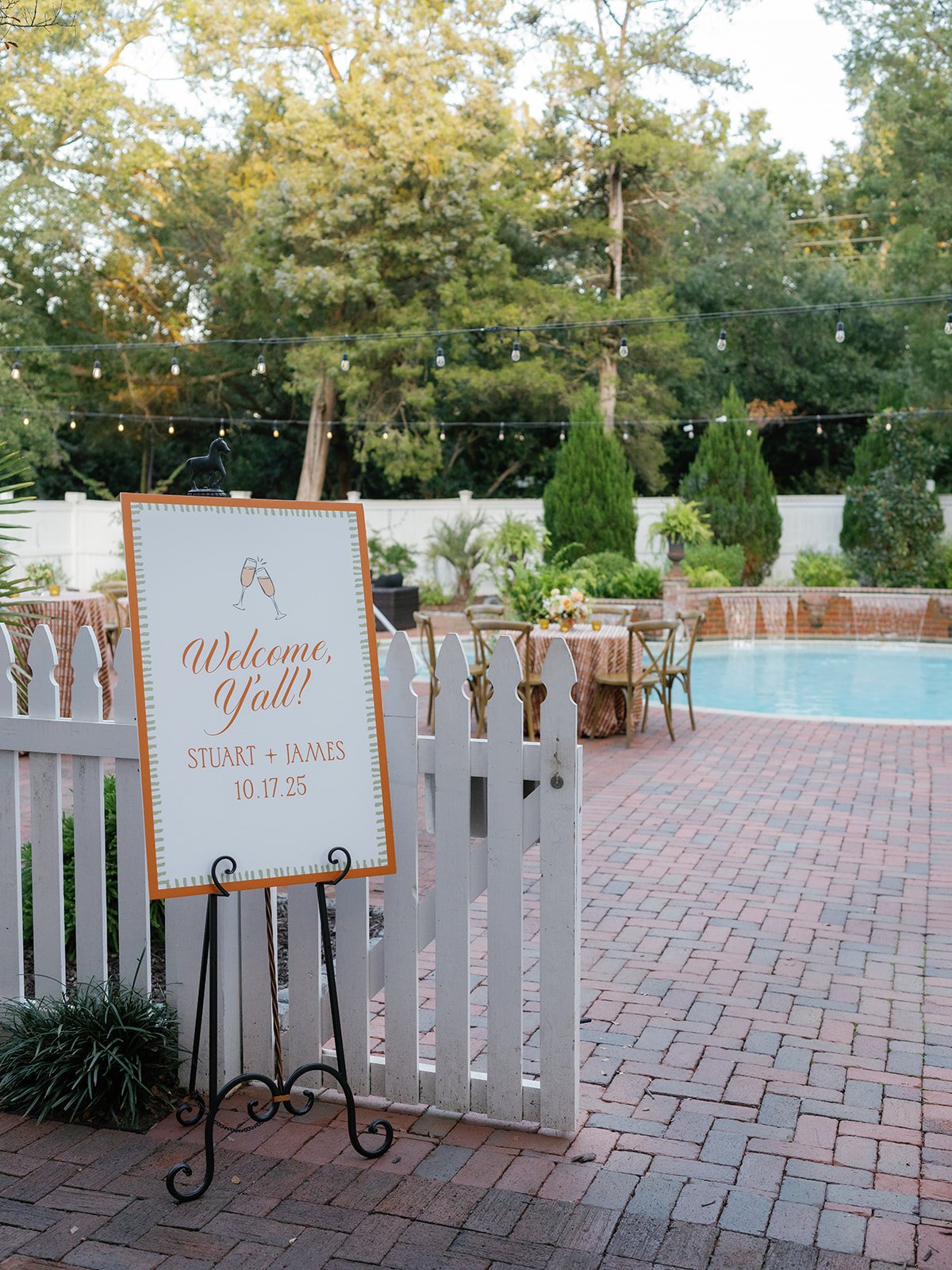 Poolside wedding welcome sign reading "Welcome, Y'all!" at the entrance to an outdoor welcome party with string lights and tables beyond.