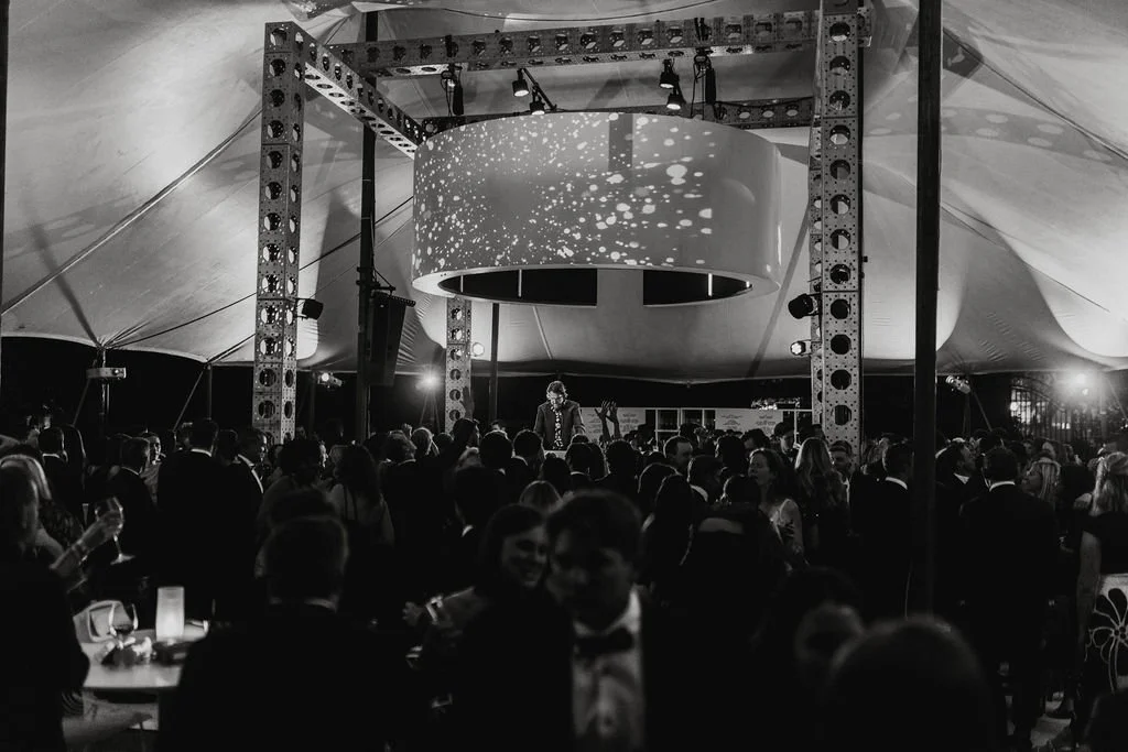 Crowded wedding dance floor gathered around a DJ booth beneath a tented reception structure with statement lighting overhead.