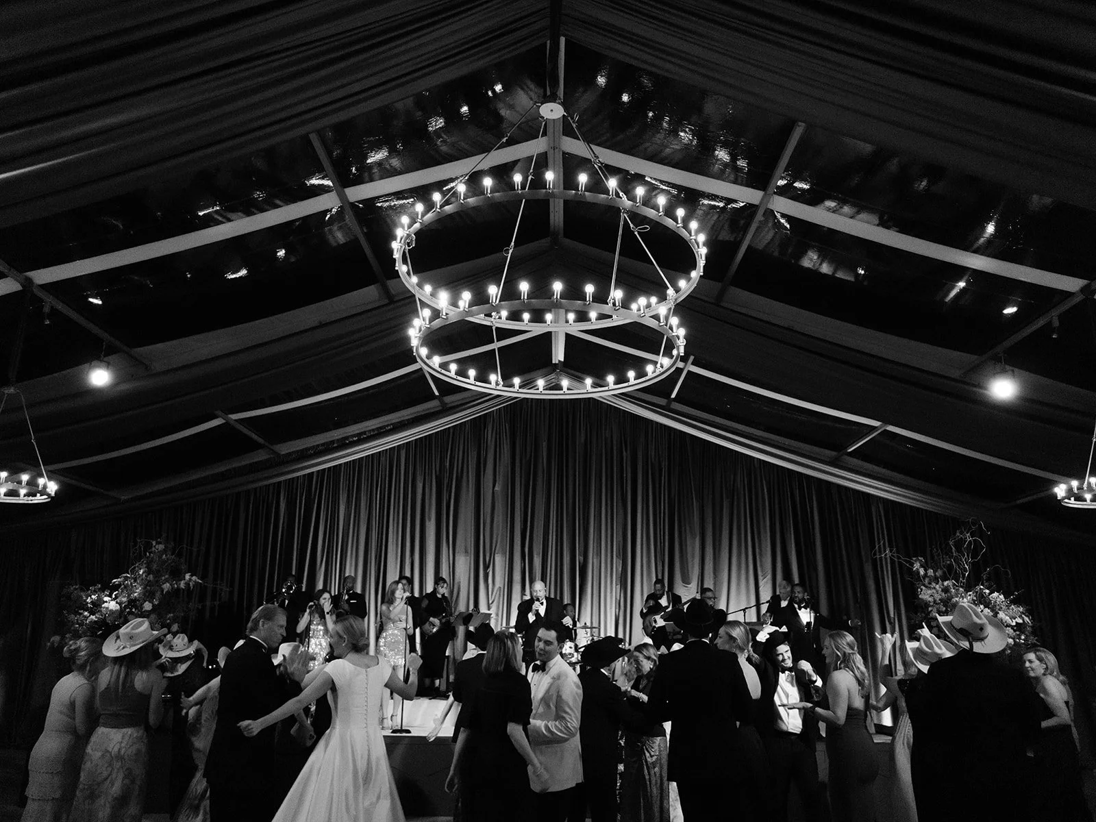 Guests dancing in front of a live wedding band inside a draped reception tent with chandelier lighting overhead.