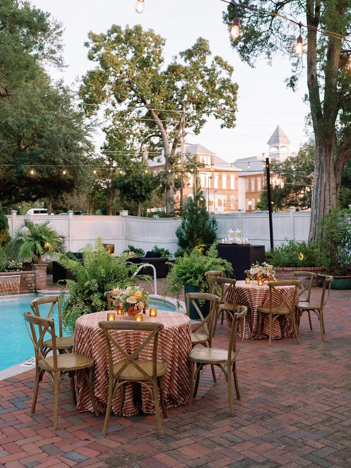 Poolside wedding welcome party seating with round tables in rust patterned linens, candlelight, café string lights, and a historic building in the background at dusk.