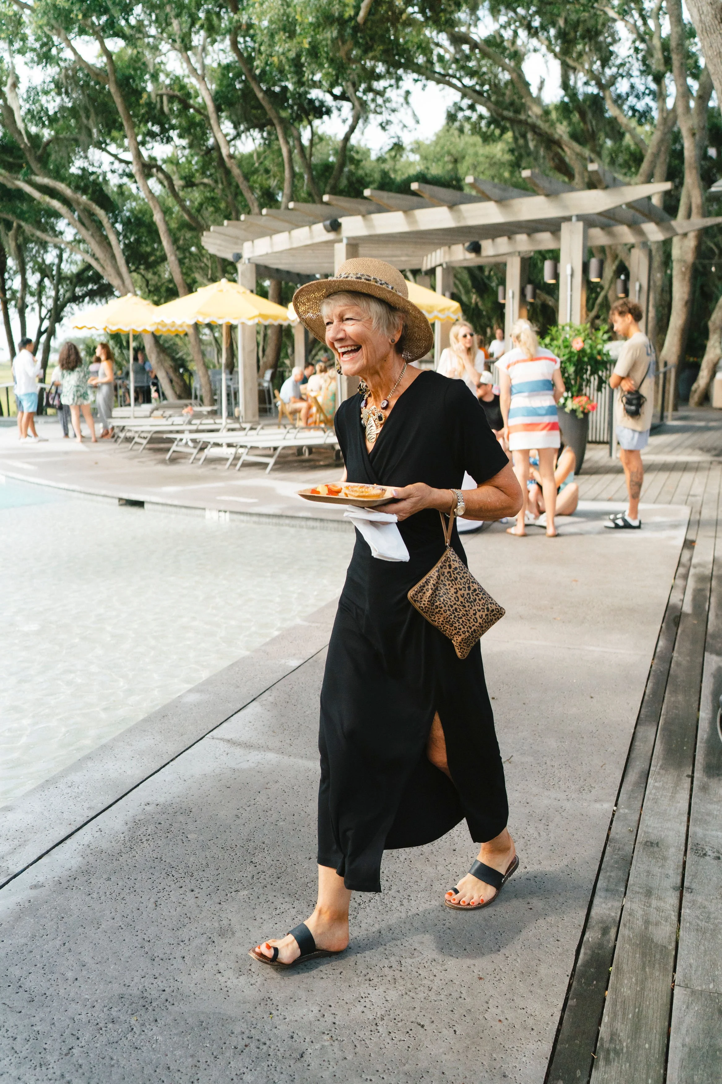 Guest in a black dress and straw hat carrying brunch by the pool during a relaxed wedding weekend farewell gathering.