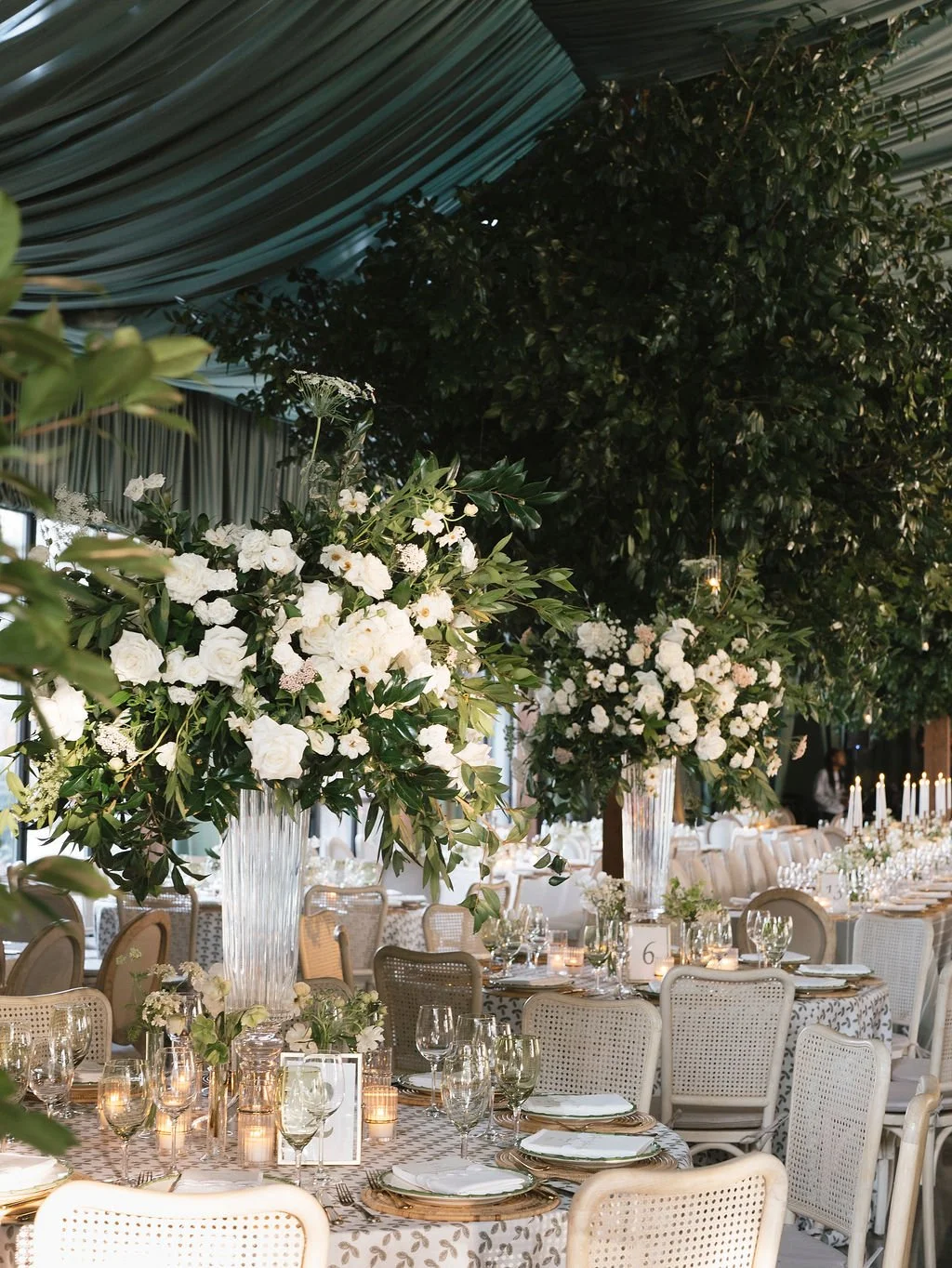 Tall garden-style floral centerpieces with white blooms and greenery in a tented wedding reception.
