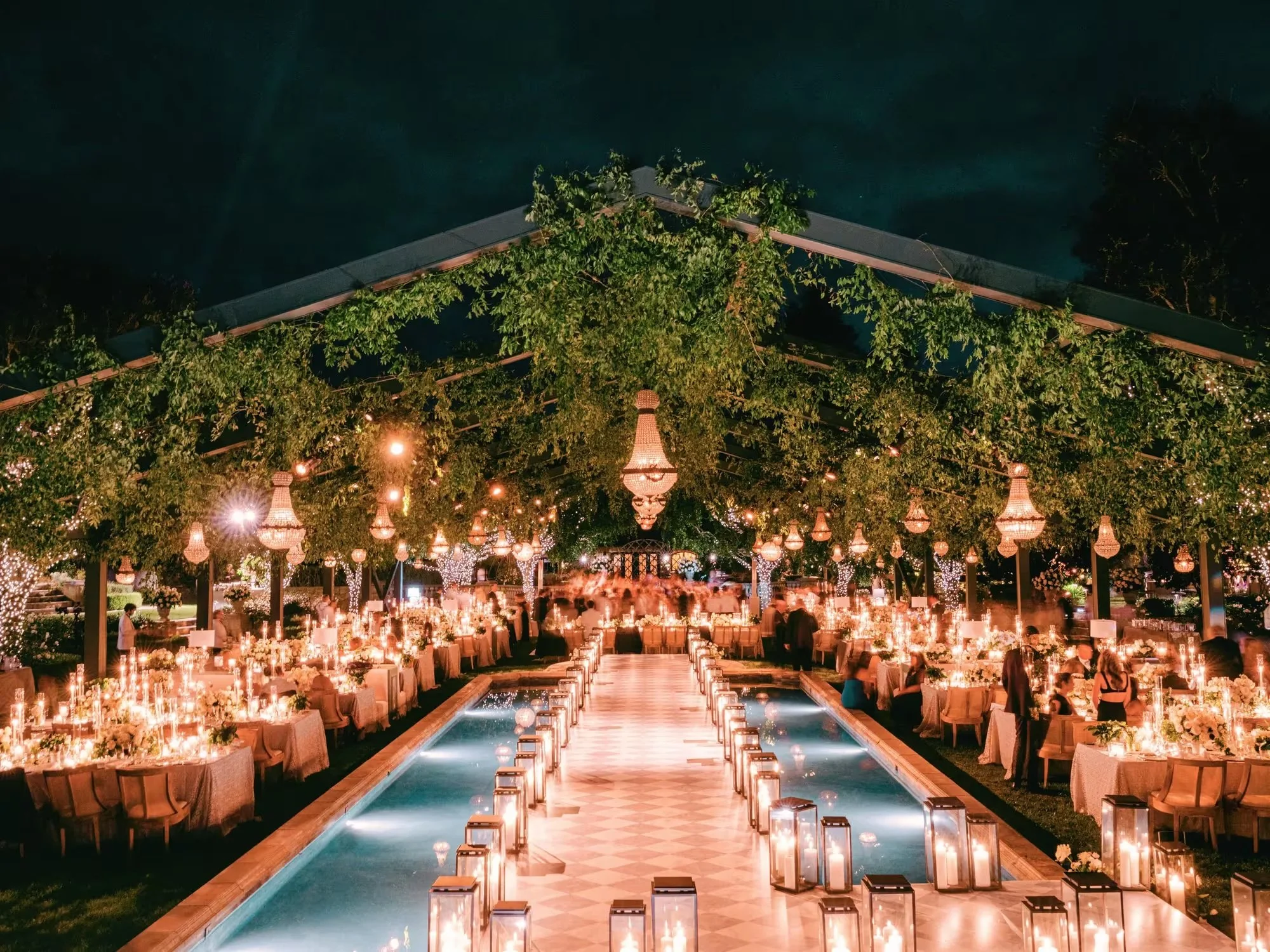 Candlelit outdoor wedding reception beside a long reflecting pool, with lanterns and chandeliers under lush greenery.