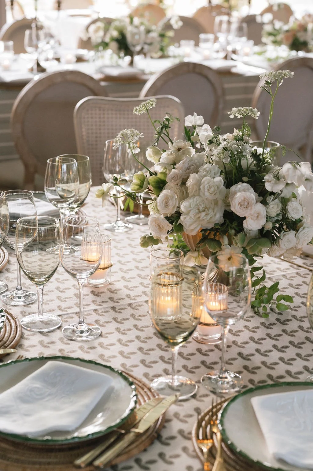 Close-up of a wedding tablescape with white garden roses, patterned linen, woven chargers, glassware, and candlelight.