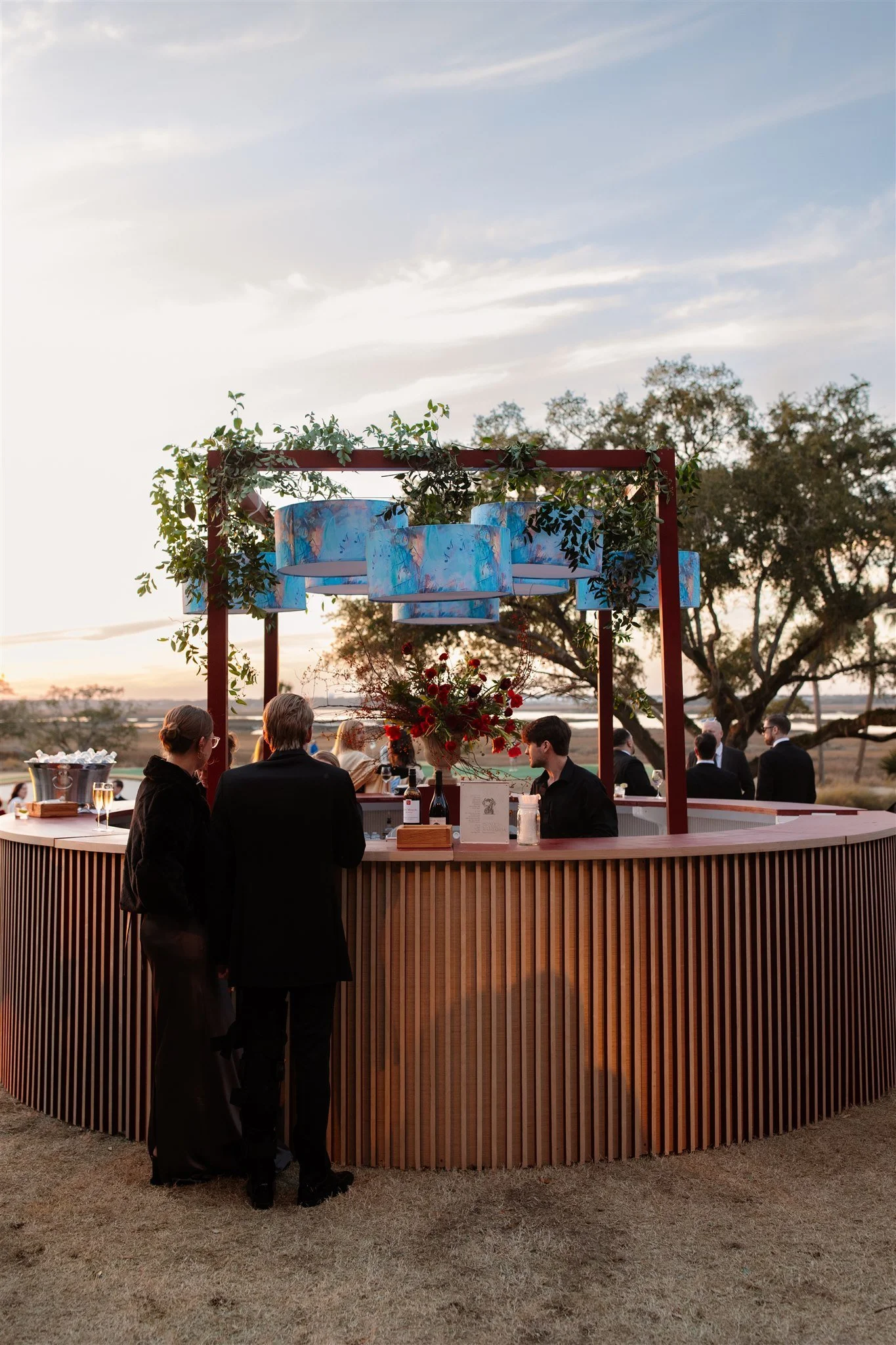 Guests order drinks at a circular wooden bar framed with greenery and blue drum lanterns during an outdoor cocktail hour.
