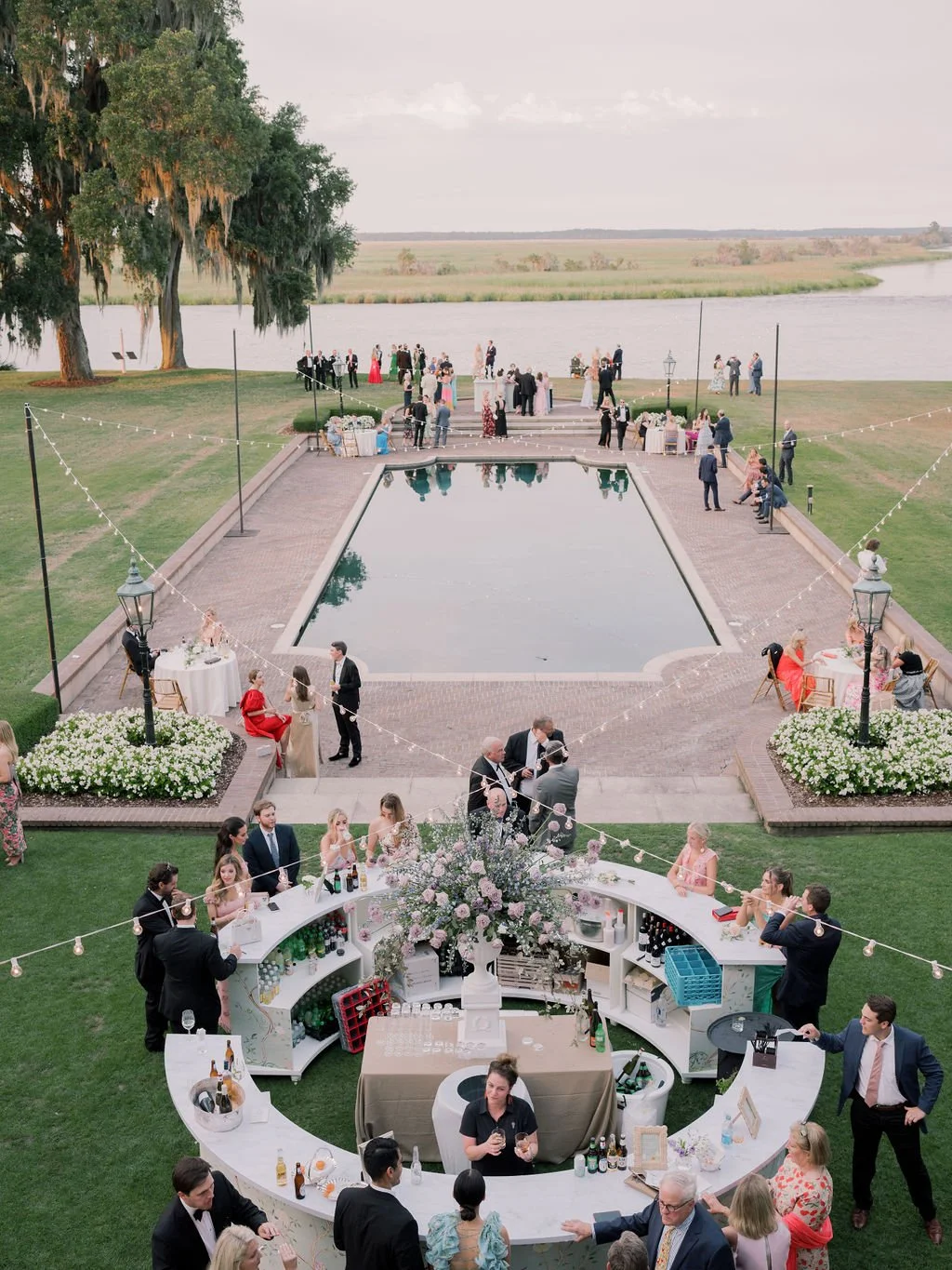 Outdoor cocktail hour at a Southern estate wedding venue with a reflecting pool, waterfront views, string lights, and guests gathered on the lawn.