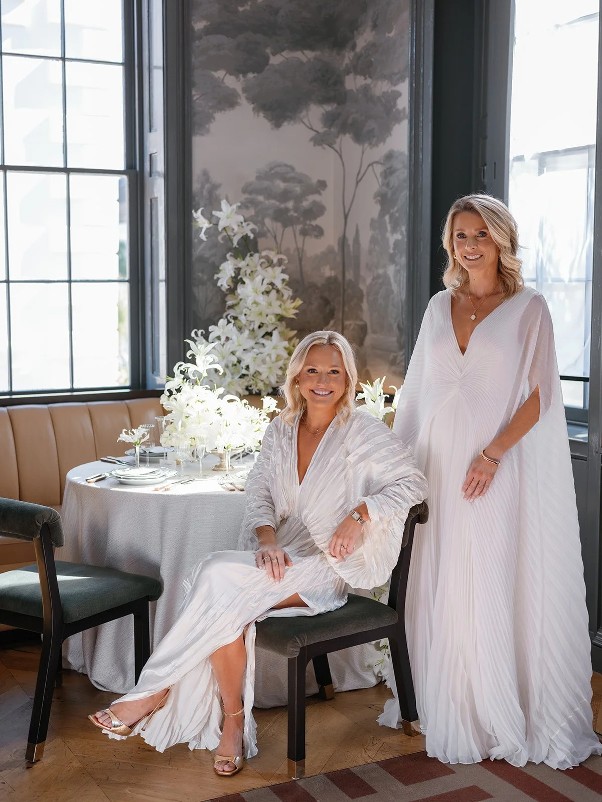 Two women in pleated white gowns pose beside a round table layered with white lilies, patterned china, and soft daylight in an elegant editorial interior.