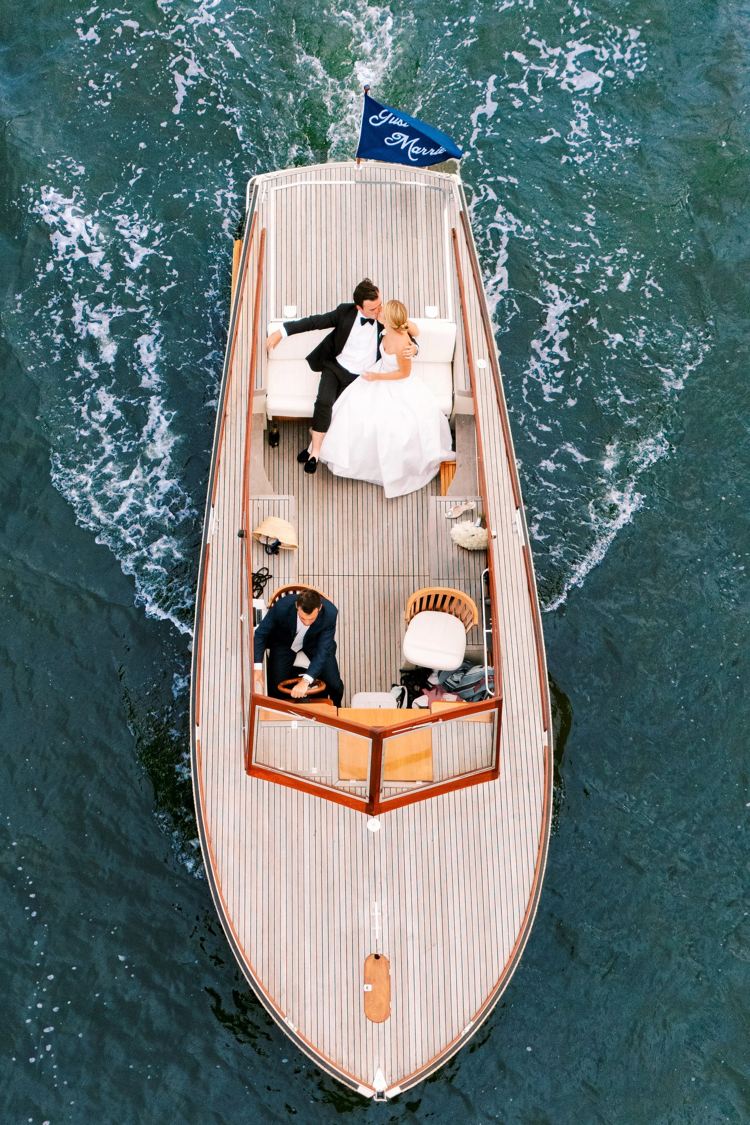 Overhead view of a couple in formalwear embracing on a classic wooden boat with a “Just Married” flag, cutting through deep blue water.