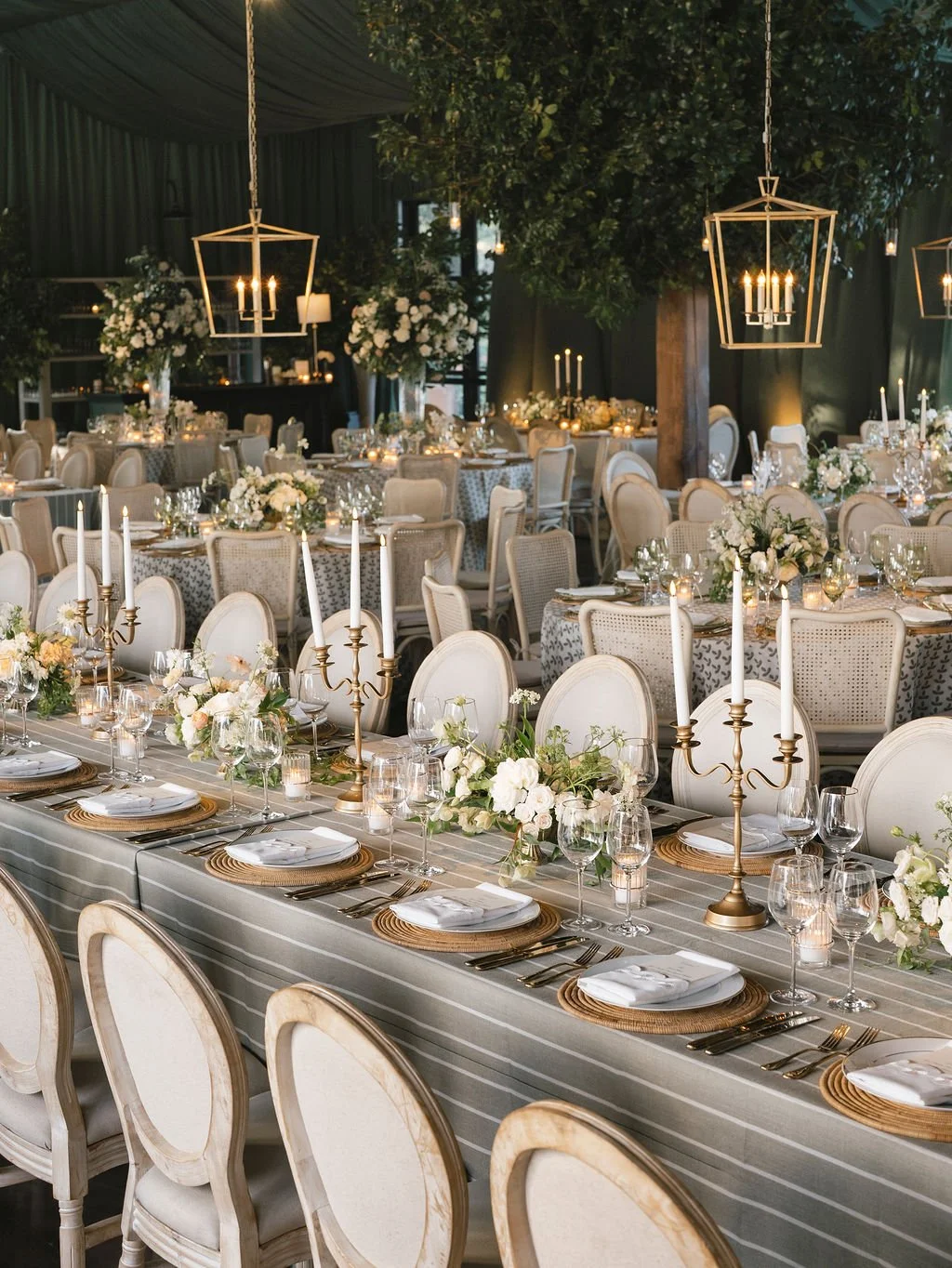 Refined reception tablescape with striped greige linens, ivory chairs, gold candlesticks, and soft candlelight under a canopy of greenery and lanterns, designed by Reagan Events.