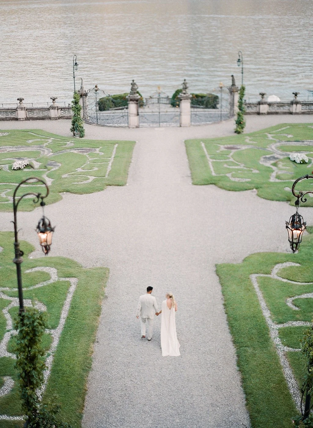 Newlywed couple walking hand in hand through the formal lakefront gardens of an Italian villa overlooking Lake Como.