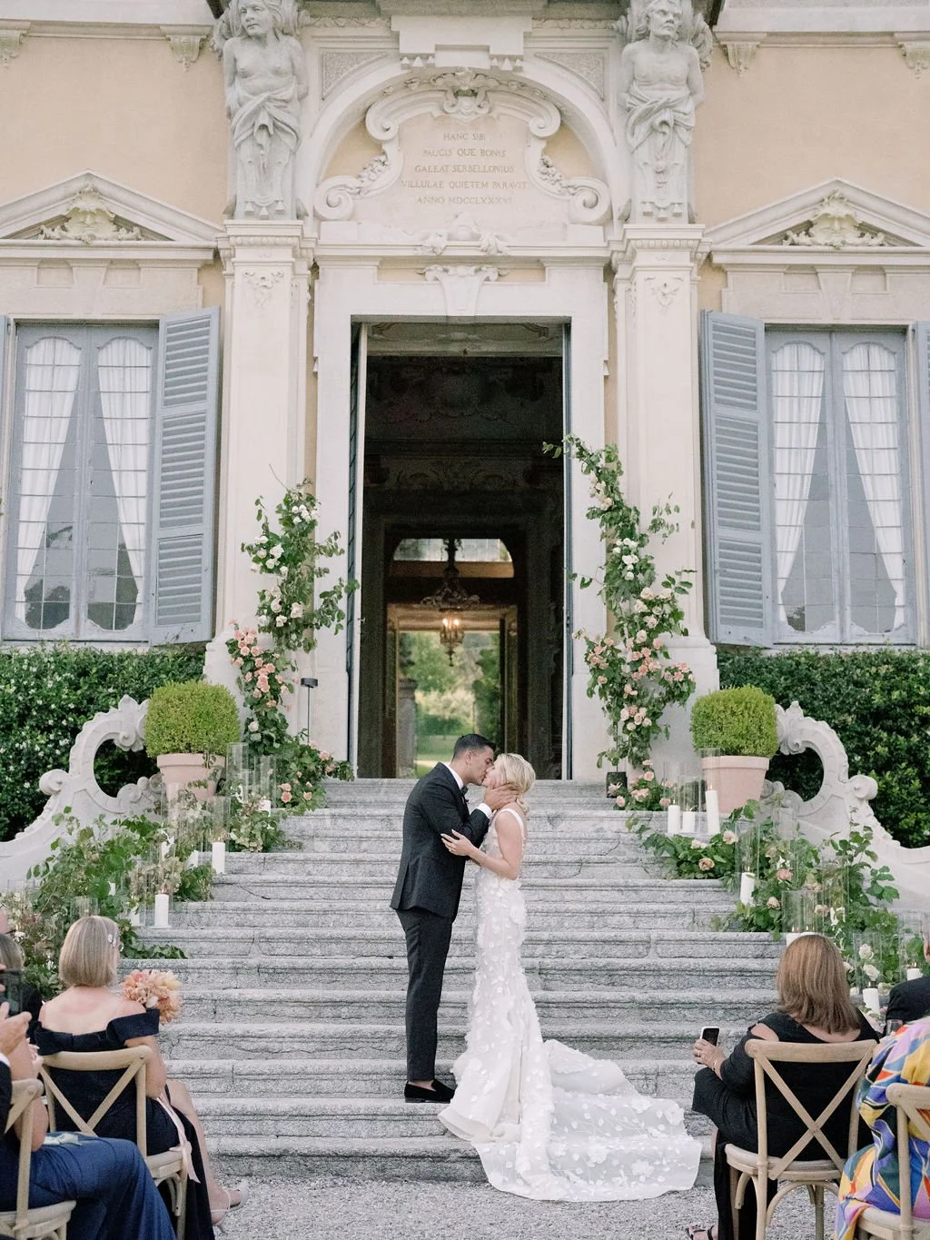 A bride and groom share a kiss on the stone steps of a historic Lake Como villa, surrounded by floral details and guests during an outdoor ceremony.v