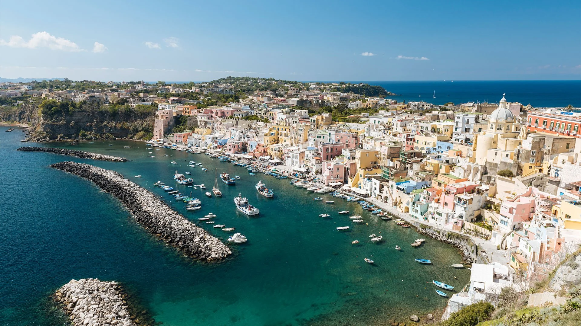 An aerial view of Procida’s harbor with pastel waterfront buildings, small boats in clear blue water, and the open sea beyond.