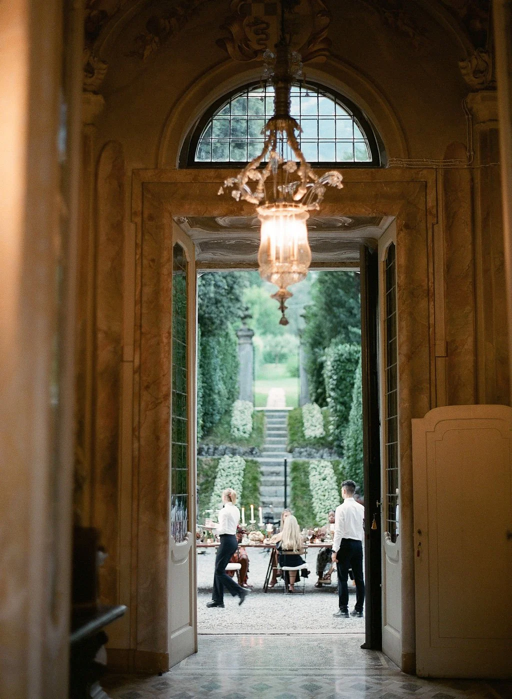 View through an ornate villa doorway toward a candlelit outdoor dinner in a formal Italian garden with guests and servers.