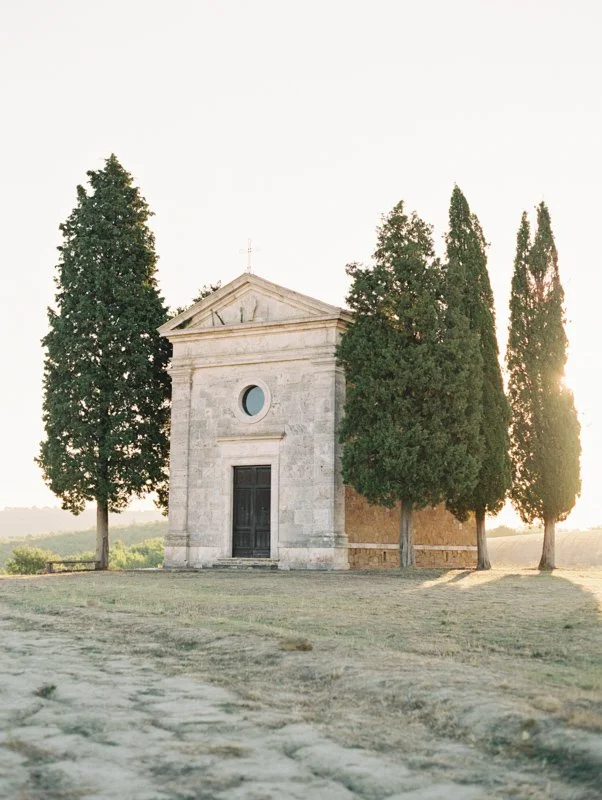 Historic stone chapel in Tuscany framed by cypress trees and soft evening light for an Italy destination wedding setting.