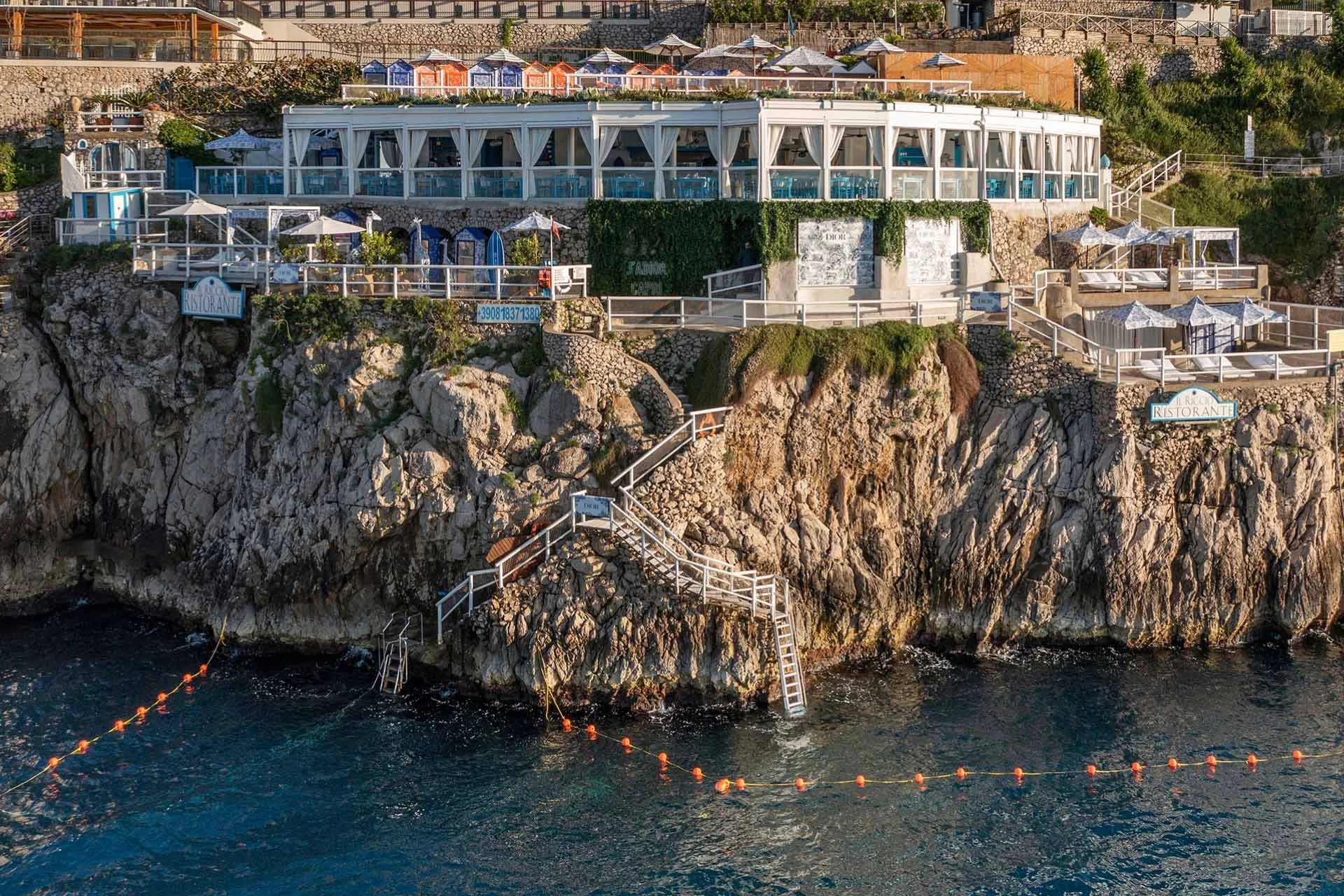 Il Riccio Restaurant and Beach Club perched on Capri’s cliffs above the water, with white cabanas, terraces, and stone steps down to the sea.