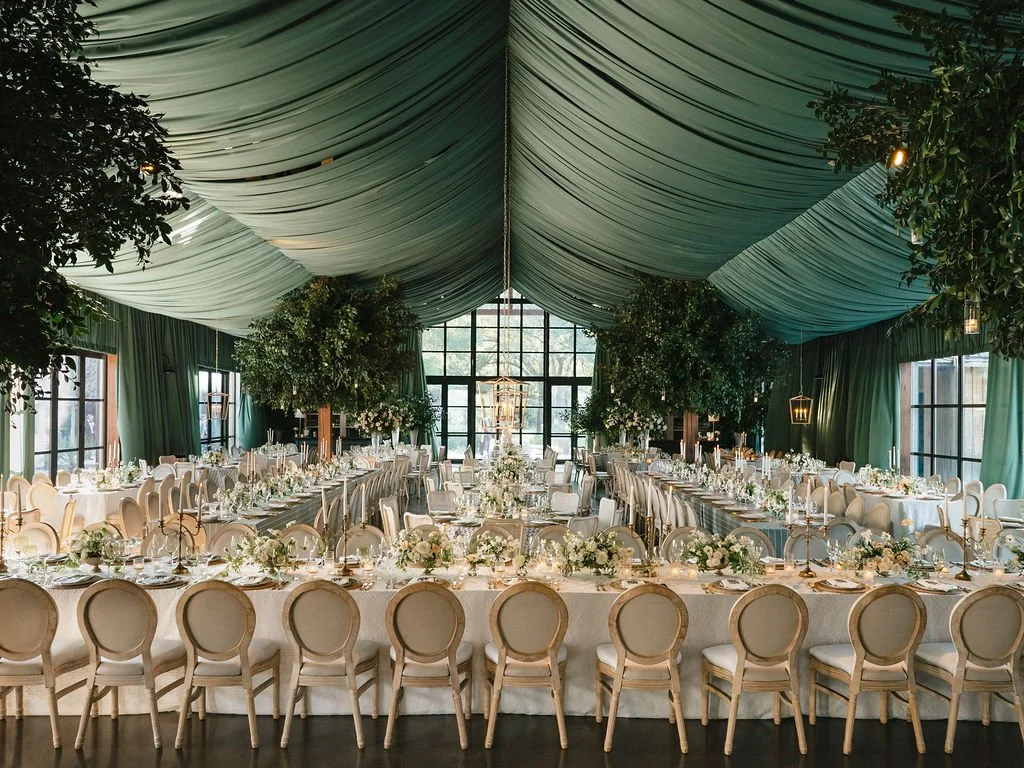 Elegant wedding reception beneath sweeping green ceiling draping with long dinner tables, candlelight, white florals, and mature trees incorporated into the design