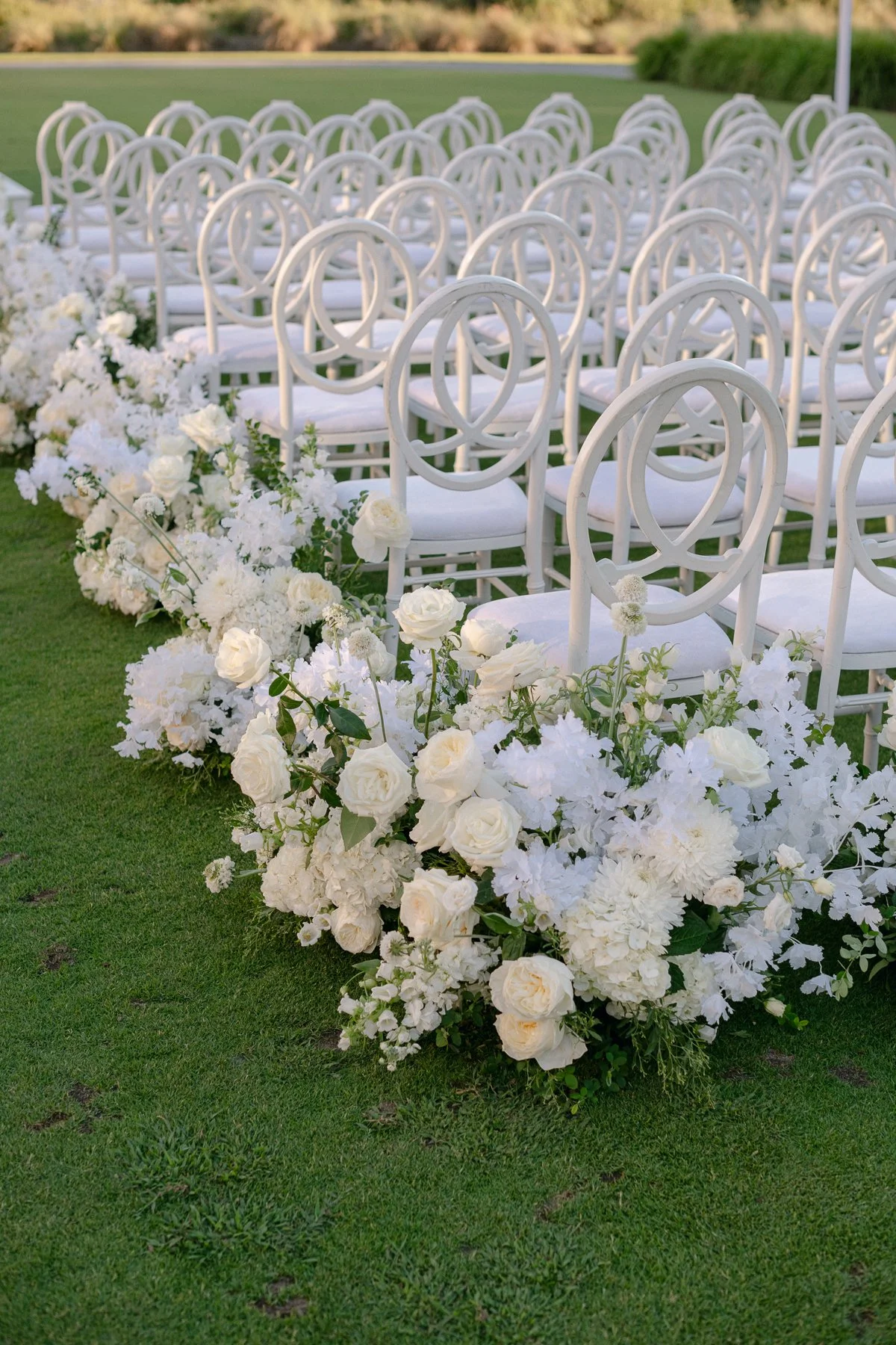 Rows of white loop-back chairs on a green lawn with a low white floral aisle design for a ceremony at The Sanctuary at Kiawah Island.