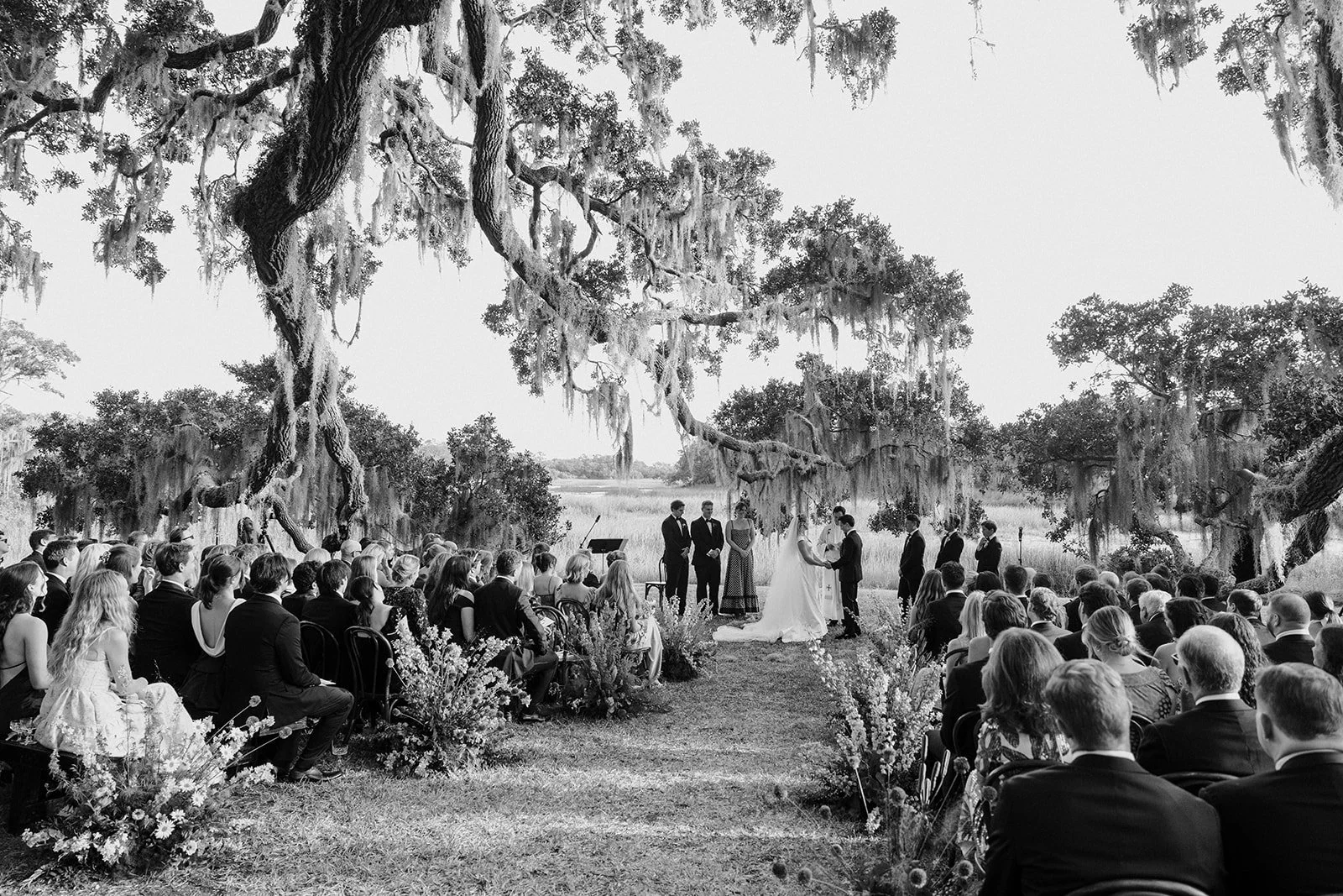 Black-and-white photograph of a Kiawah Island wedding ceremony beneath sweeping live oaks and Spanish moss overlooking the marsh.