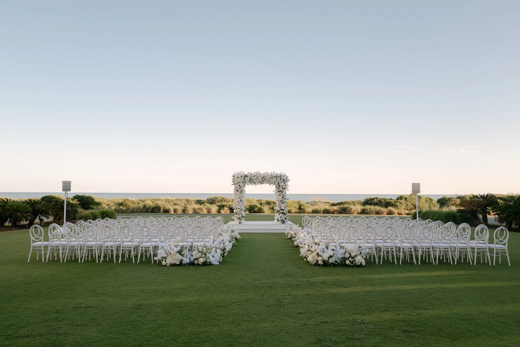 Oceanfront wedding ceremony at The Sanctuary on Kiawah Island with white chairs, a floral aisle, and a white flower-covered ceremony arch.