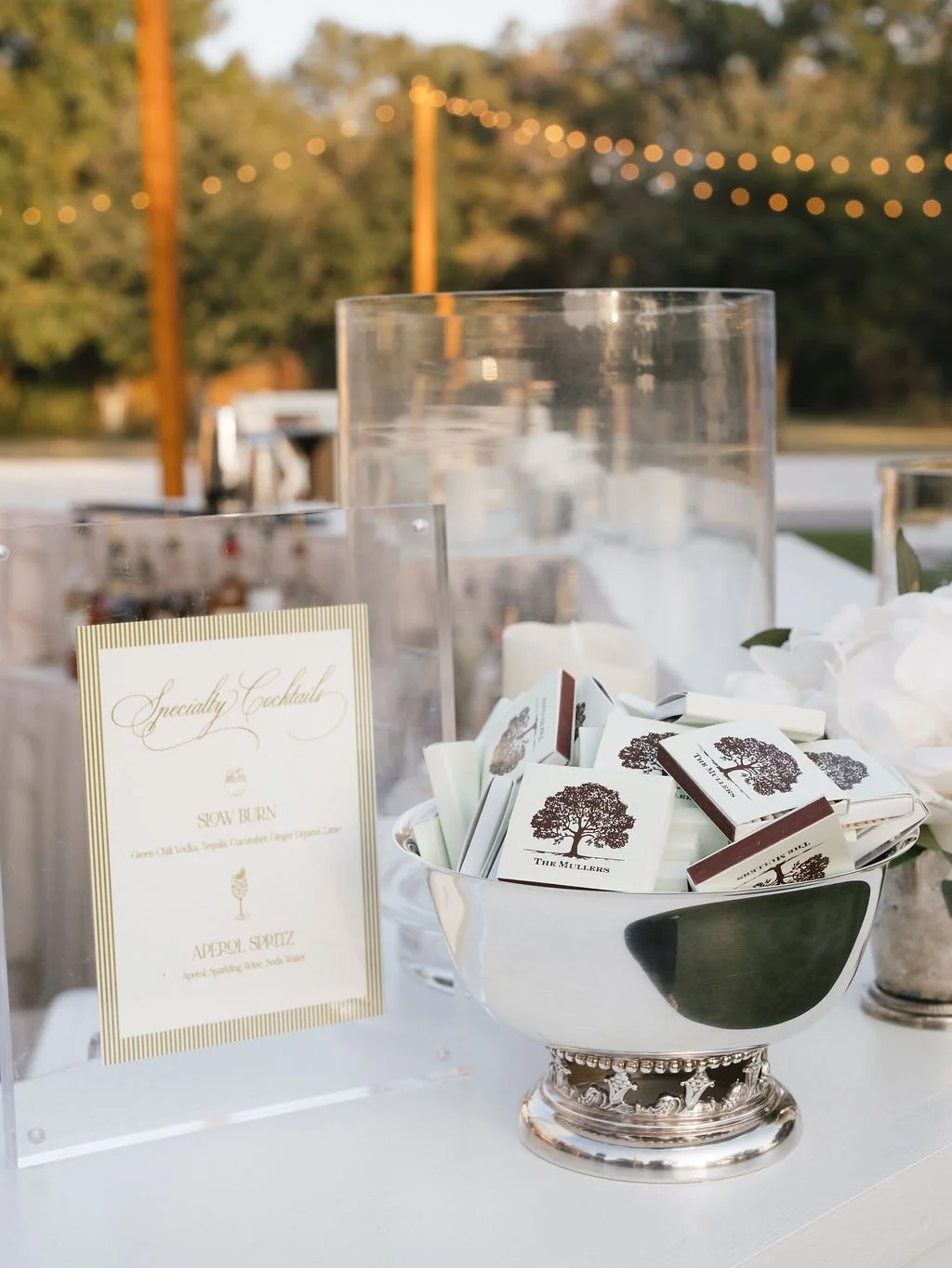Elegant specialty cocktail menu with custom matchbooks displayed in a silver bowl at an outdoor Charleston wedding bar, captured during golden hour.